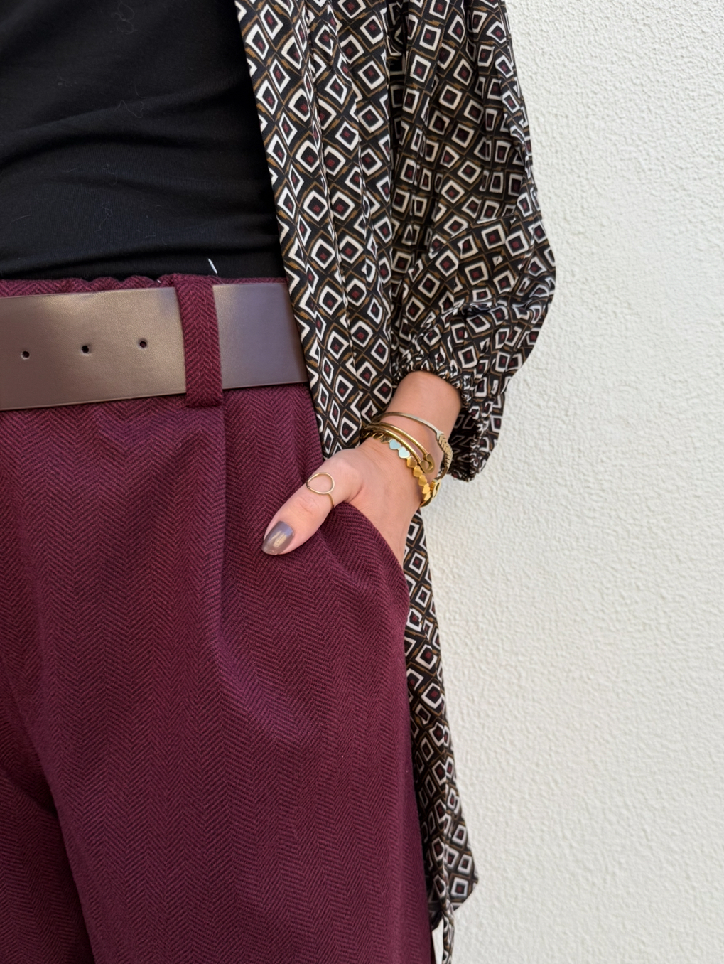 pessoa com calças bordeaux, cinto castanho, blusa preta e casaco de padrão geométrico com pulseiras e anel dourados
