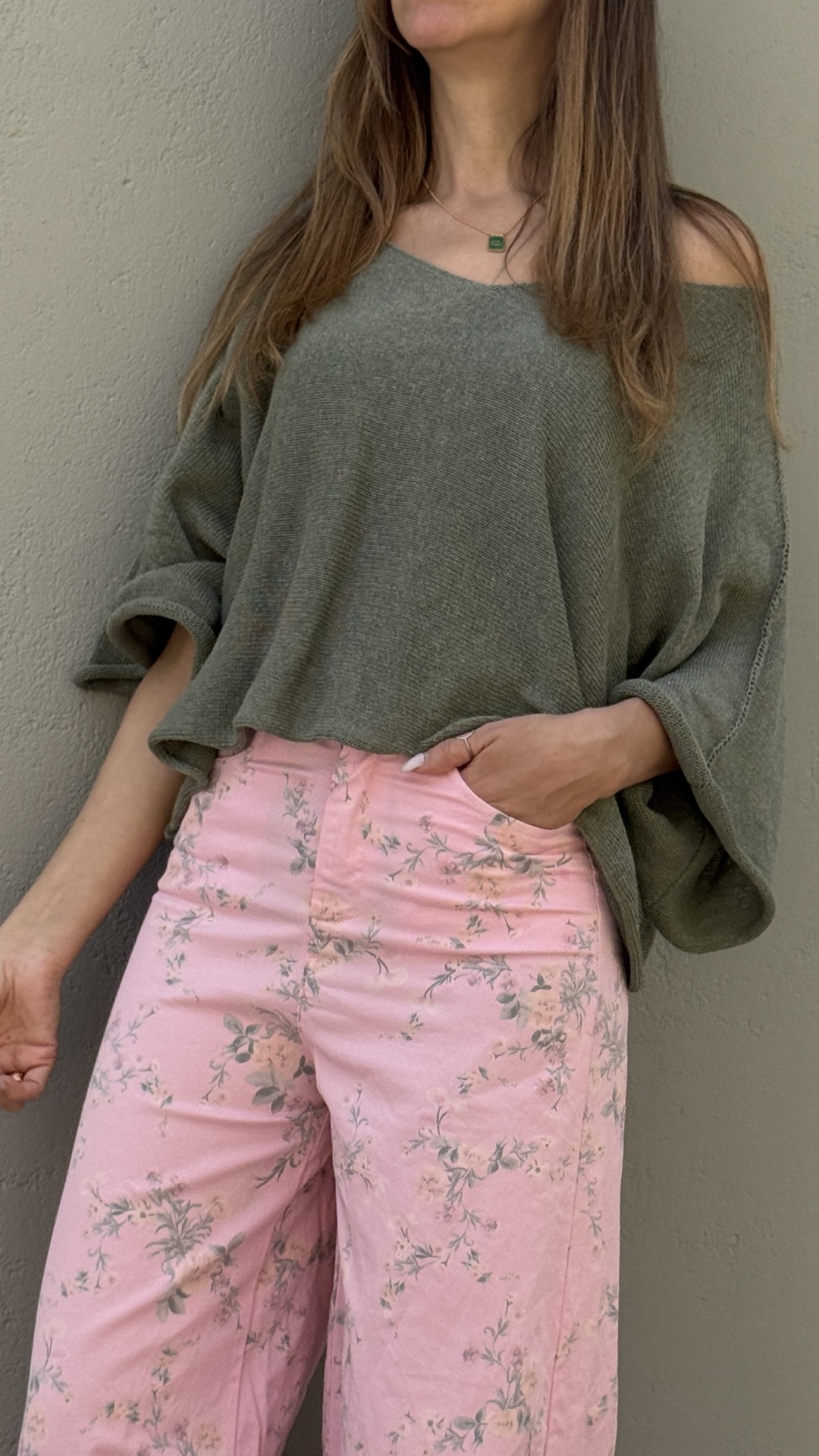 Mulher com blusa verde e calças cor-de-rosa com padrão floral junto a parede cinza.