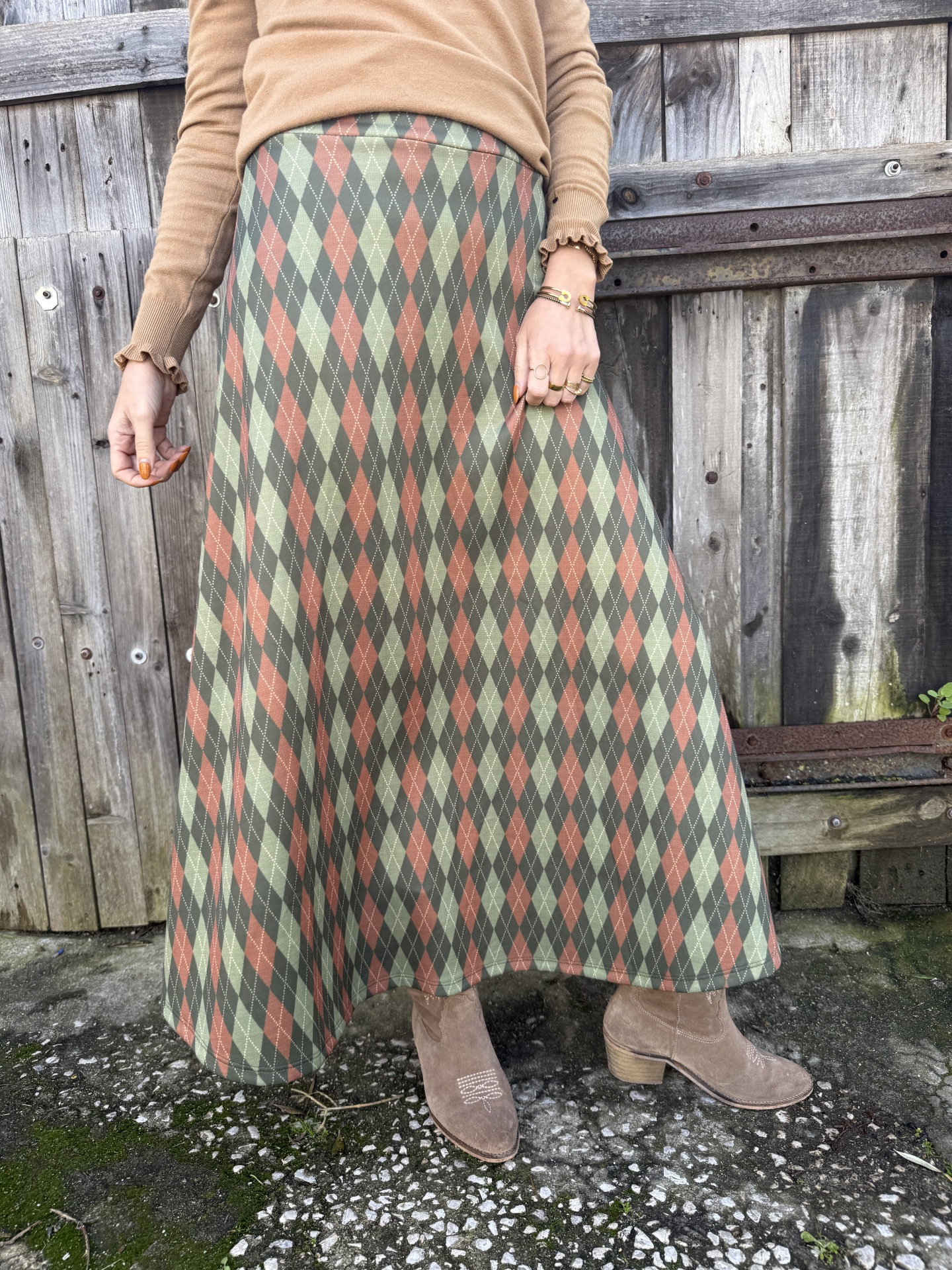 Saia longa xadrez verde laranja cinza com camisola castanha e botas camurça castanhas junto a vedação de madeira.