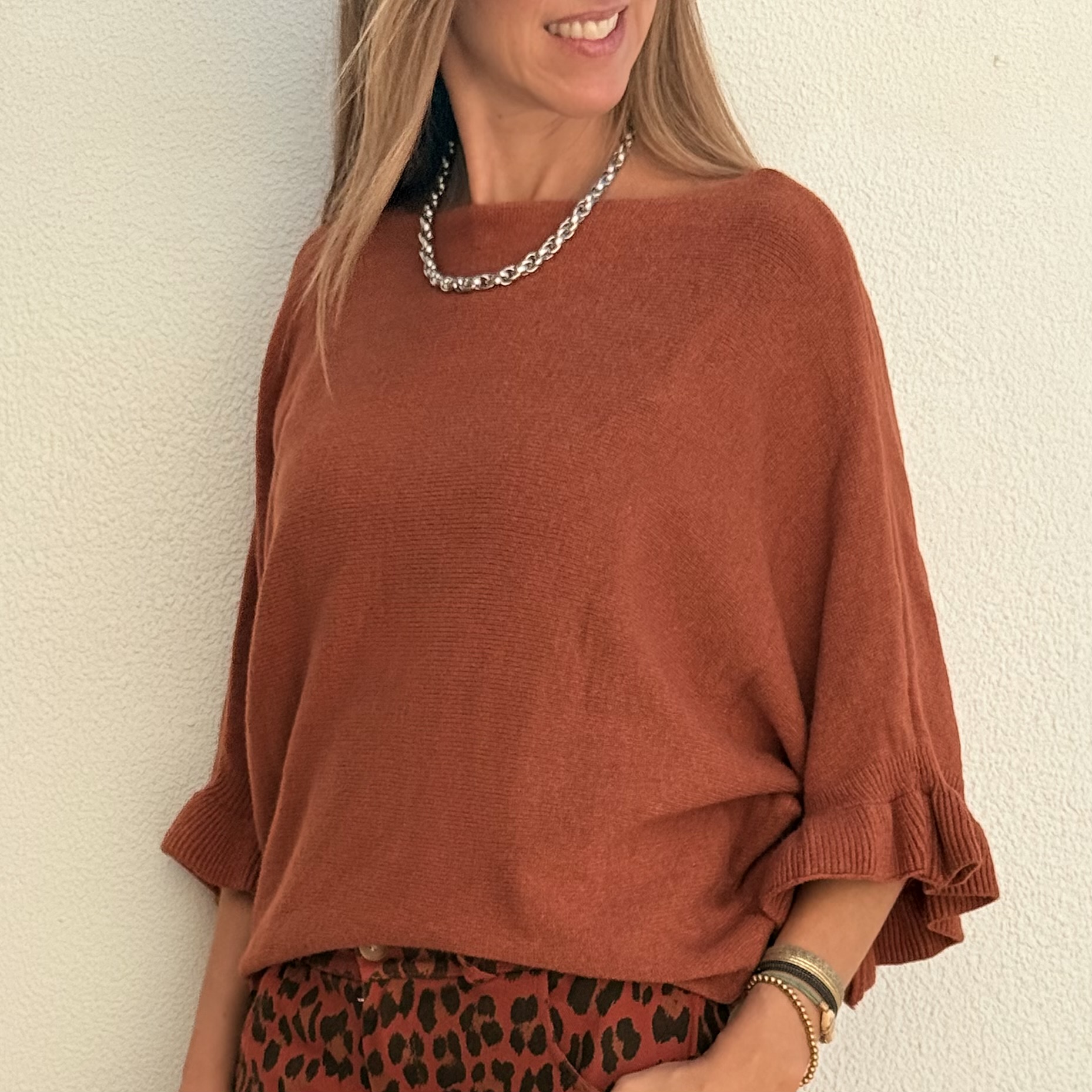 Mulher com camisola castanha de malha e colar prateado numa parede branca