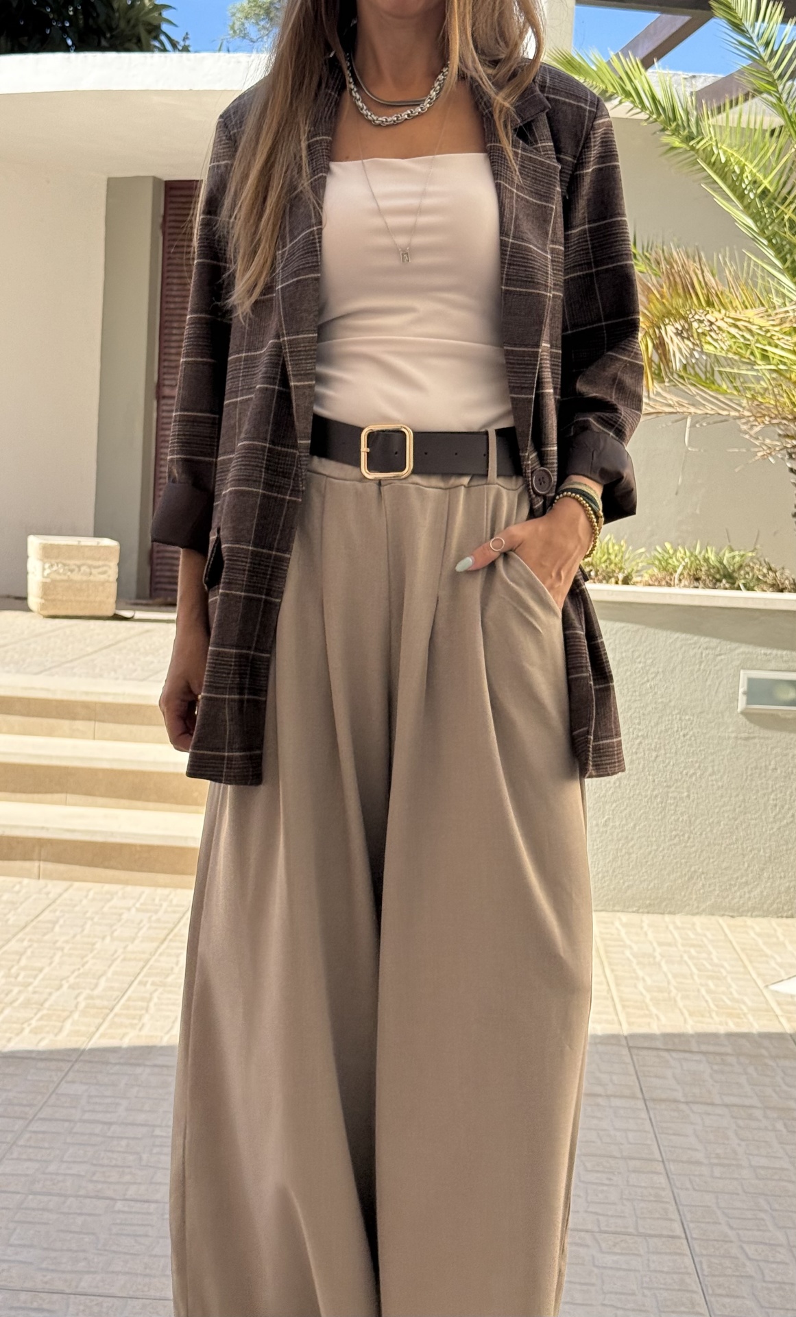 Look com casaco xadrez castanho, top branco e calças largas bege com cinto preto em ambiente exterior