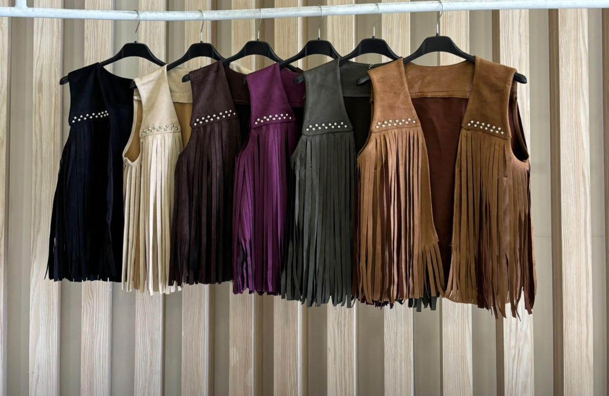 Seis coletes de franjas em camurça com tachas, diversas cores, pendurados em cabides