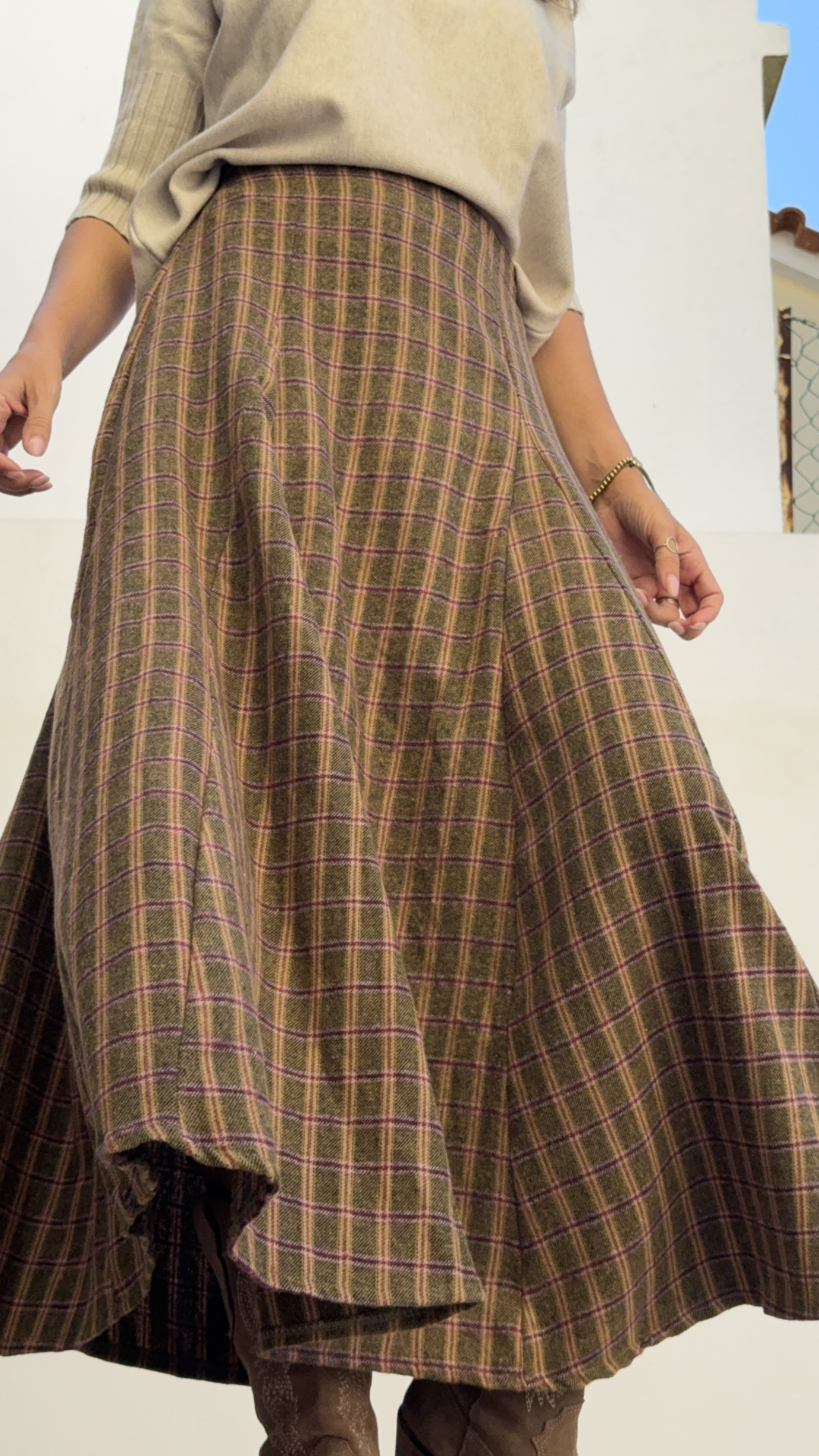 Saia midi xadrez castanha com lilás, botins castanhos e camisola bege em fundo branco e céu azul