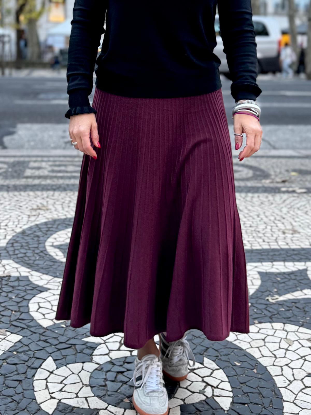 Saia midi plissada vinho e camisola preta com sapatilhas brancas numa calçada portuguesa.