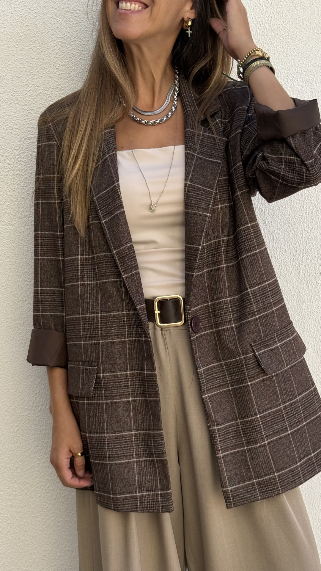 Mulher vestindo blazer xadrez castanho, camisa bege, calças largas bege e acessórios dourados e prateados