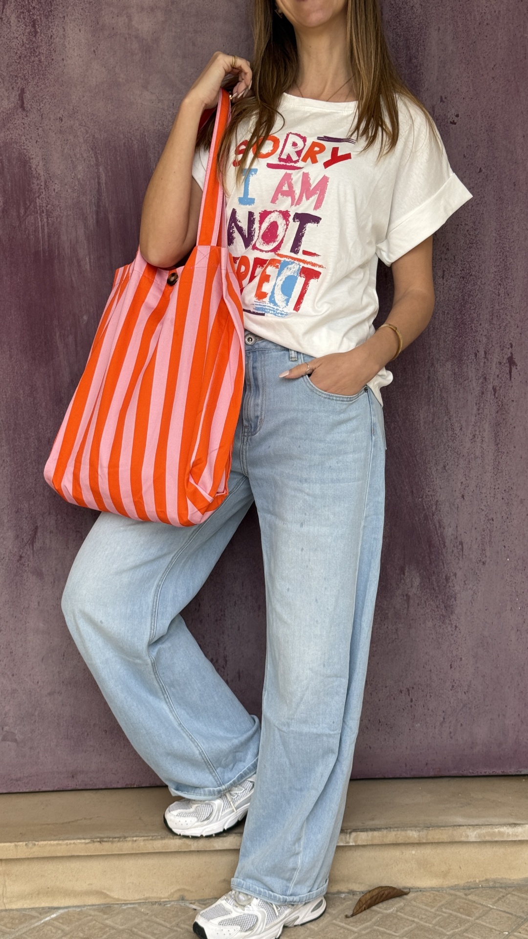 Mulher com t-shirt branca com texto colorido, calças de ganga claras, ténis brancos e mala às riscas vermelhas e rosa