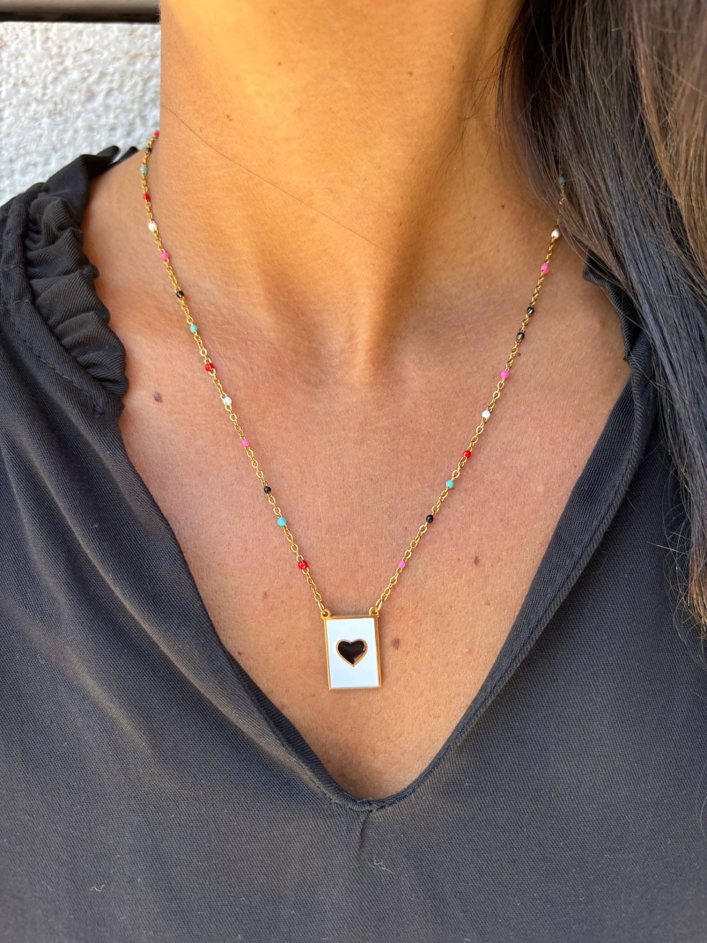 Colar dourado com pendente branco em forma de retângulo com coração recortado, usado no pescoço