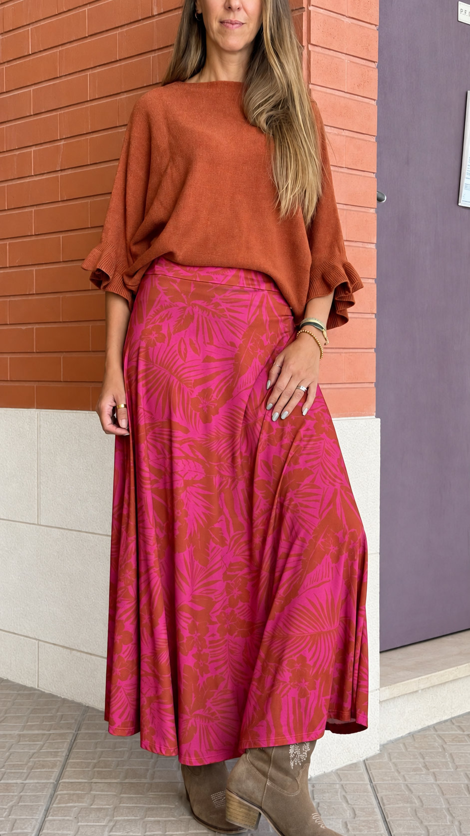 Mulher com camisola castanha, saia comprida vermelha e rosa com padrão floral, e botas castanhas.