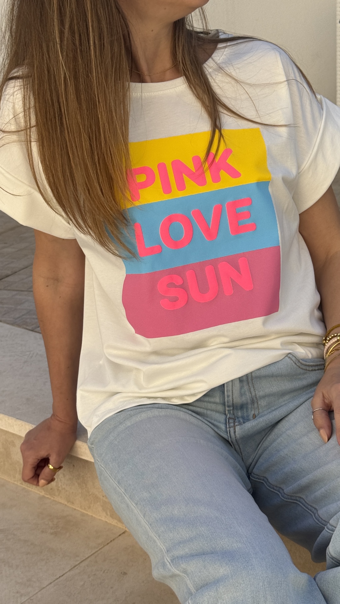 T-shirt branca PINK LOVE SUN com barras amarelas, azuis e rosas, jeans azuis claros.