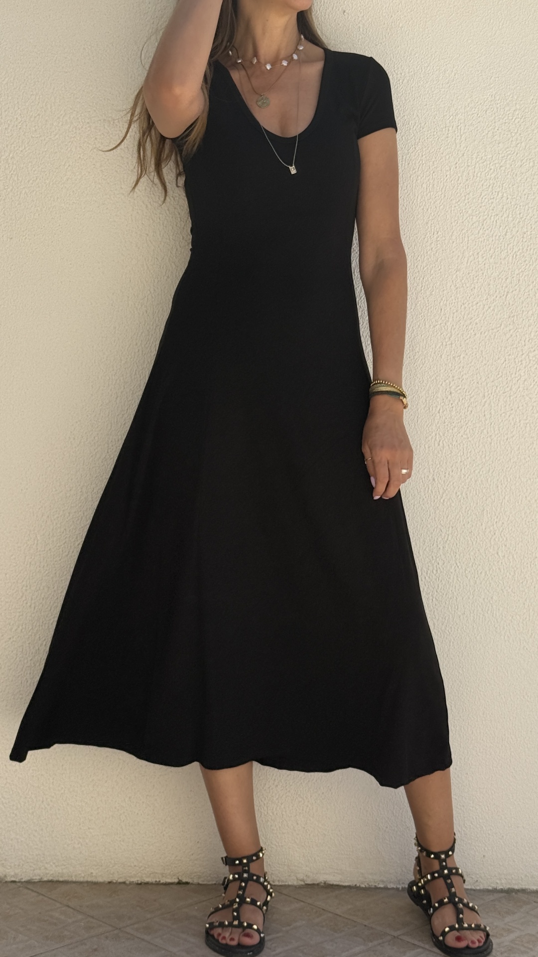 Mulher com vestido midi preto, sandálias pretas com tachas, acessórios dourados e prateados, em fundo branco.