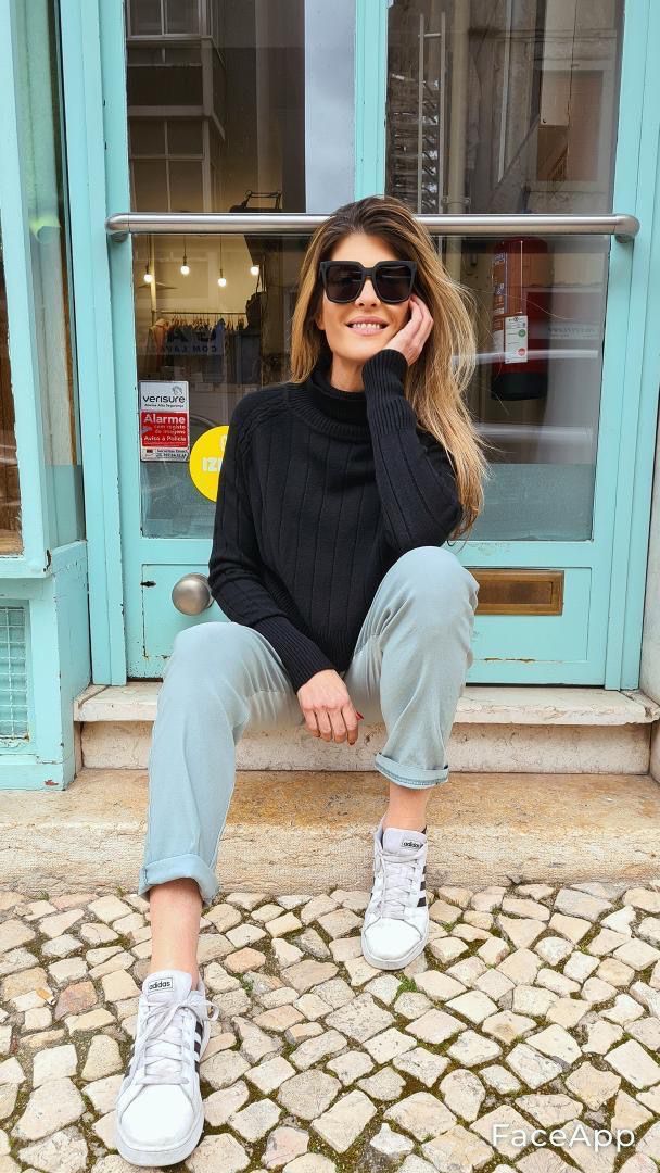 Mulher com camisola preta, calças cinza claro e ténis brancos sentada em degraus diante de porta azul clara