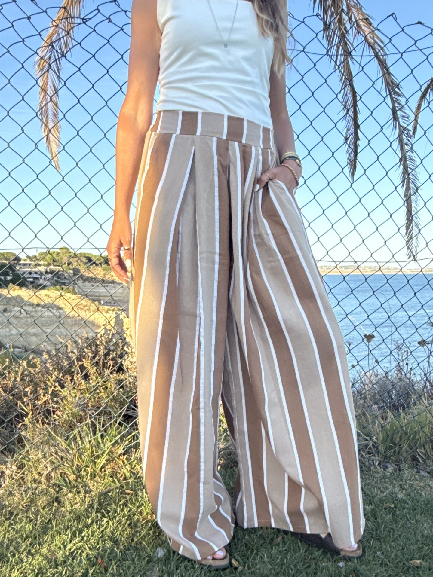 Calças largas às riscas castanhas e brancas com roupa branca e fundo costeiro