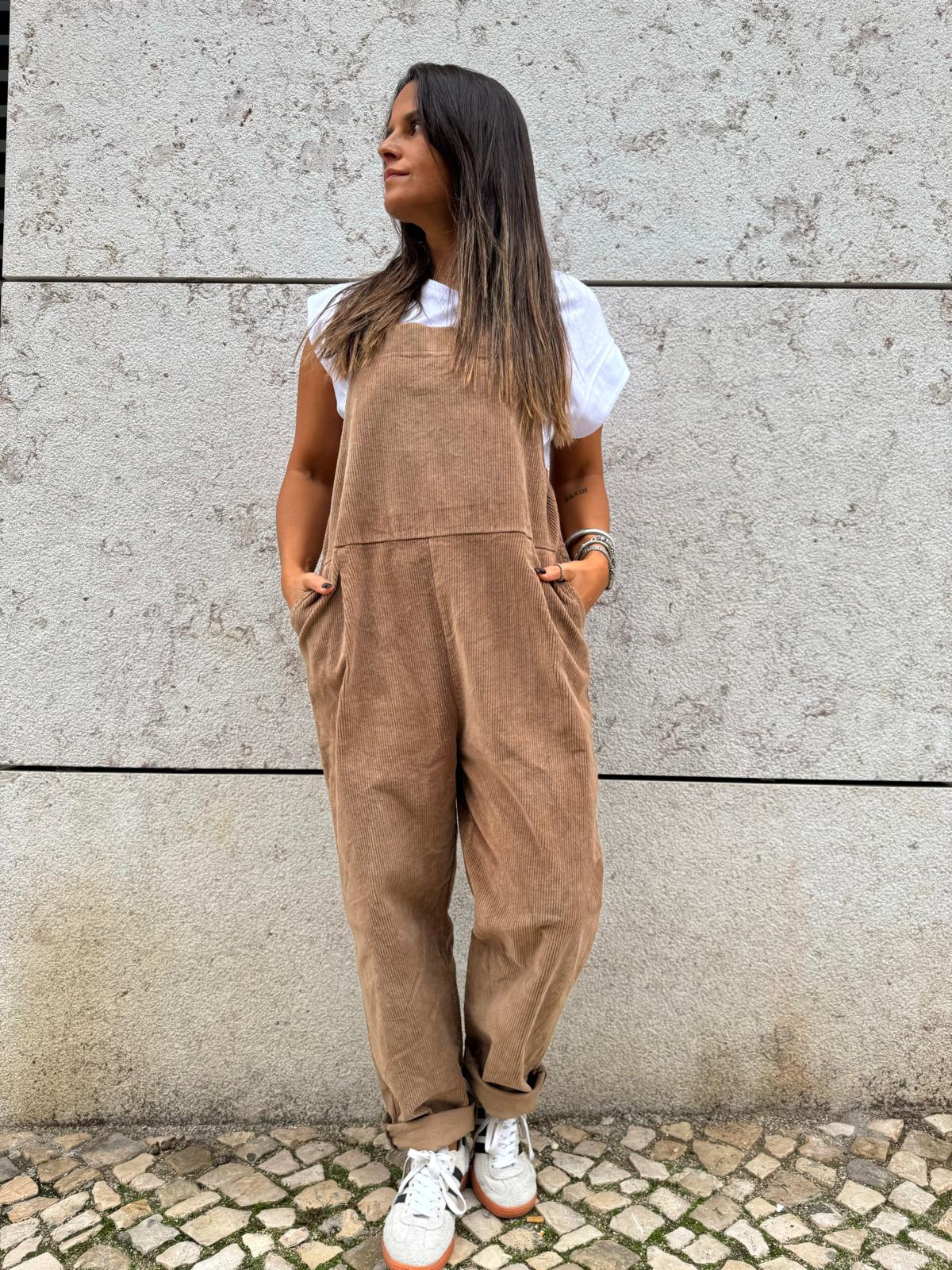 Mulher com macacão castanho claro, t-shirt branca e sapatilhas brancas em parede cinzenta
