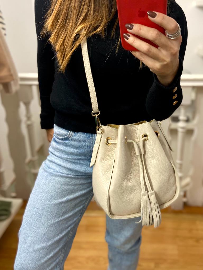 Mala branca tipo saco em pele com tassel, usada por pessoa com camisola preta e jeans azuis