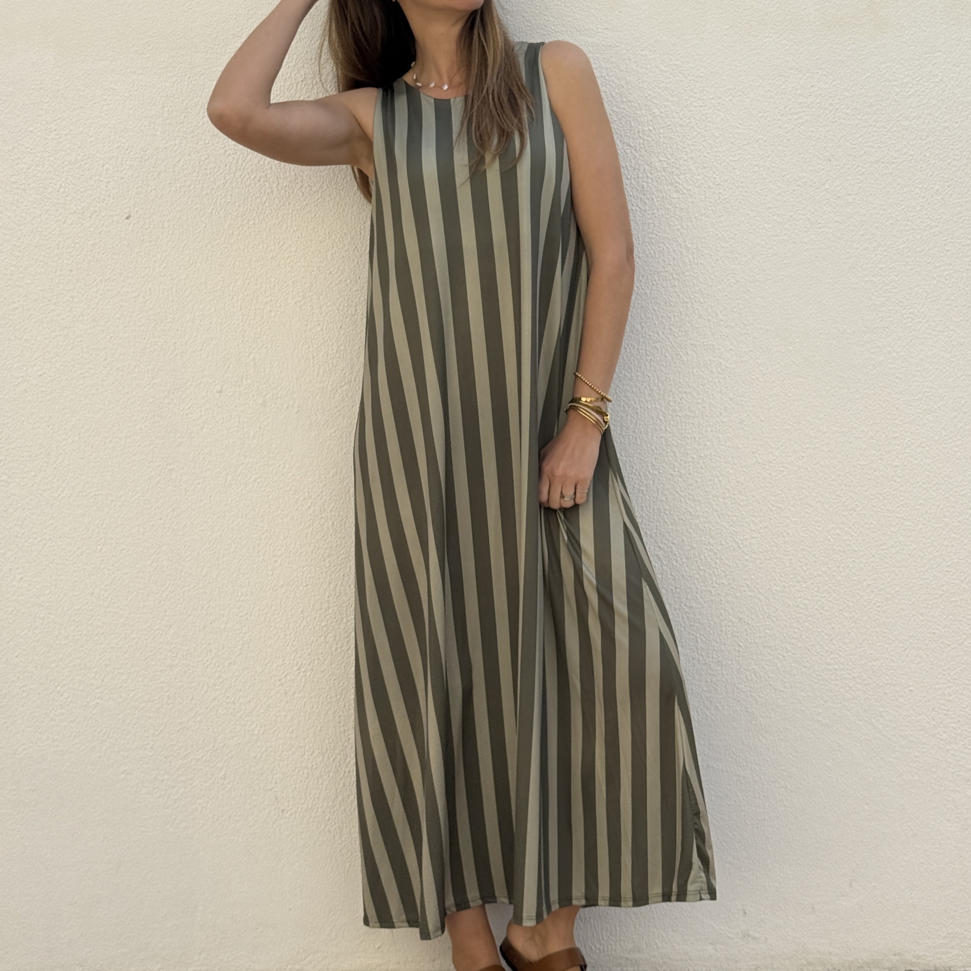 Vestido comprido listrado verde-oliva e cinzento assente em mulher com pulseiras e sandálias castanhas, fundo creme.