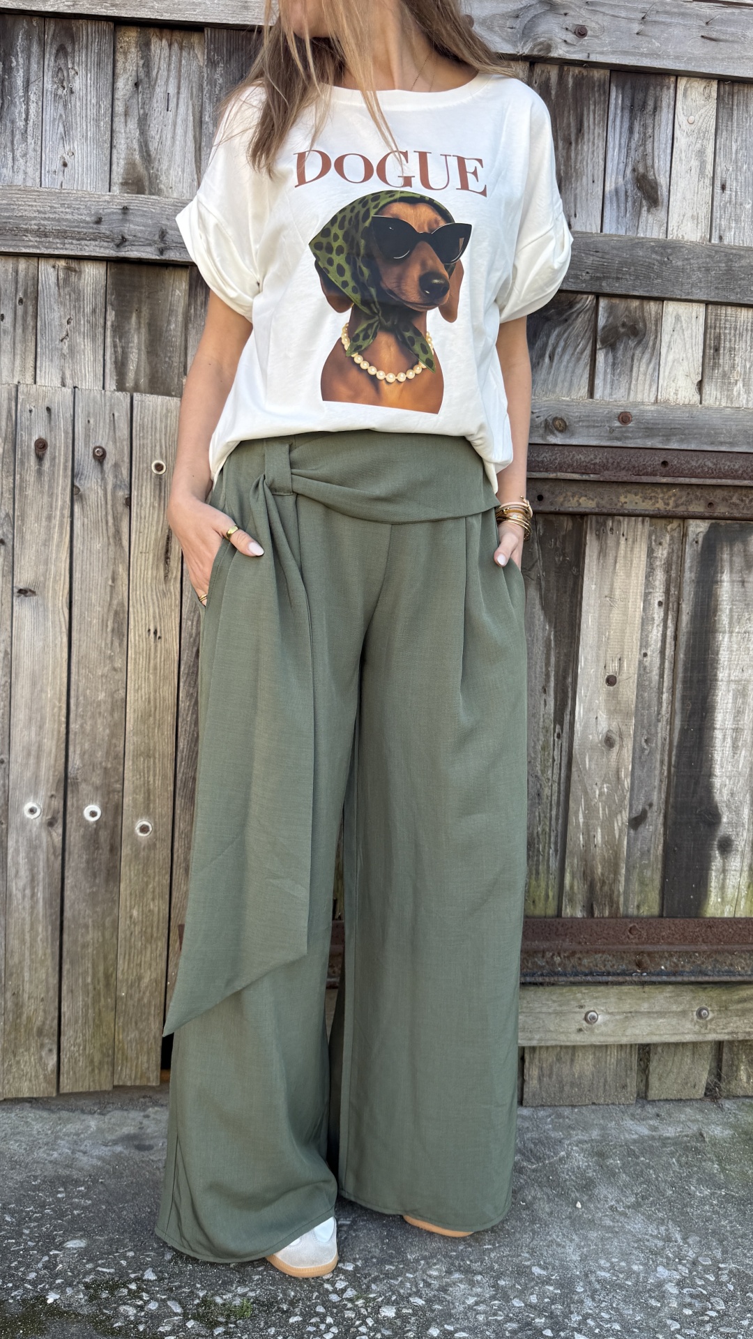 Conjunto com t-shirt branco DOGUE e calças largas verdes com laço junto a uma cerca de madeira envelhecida