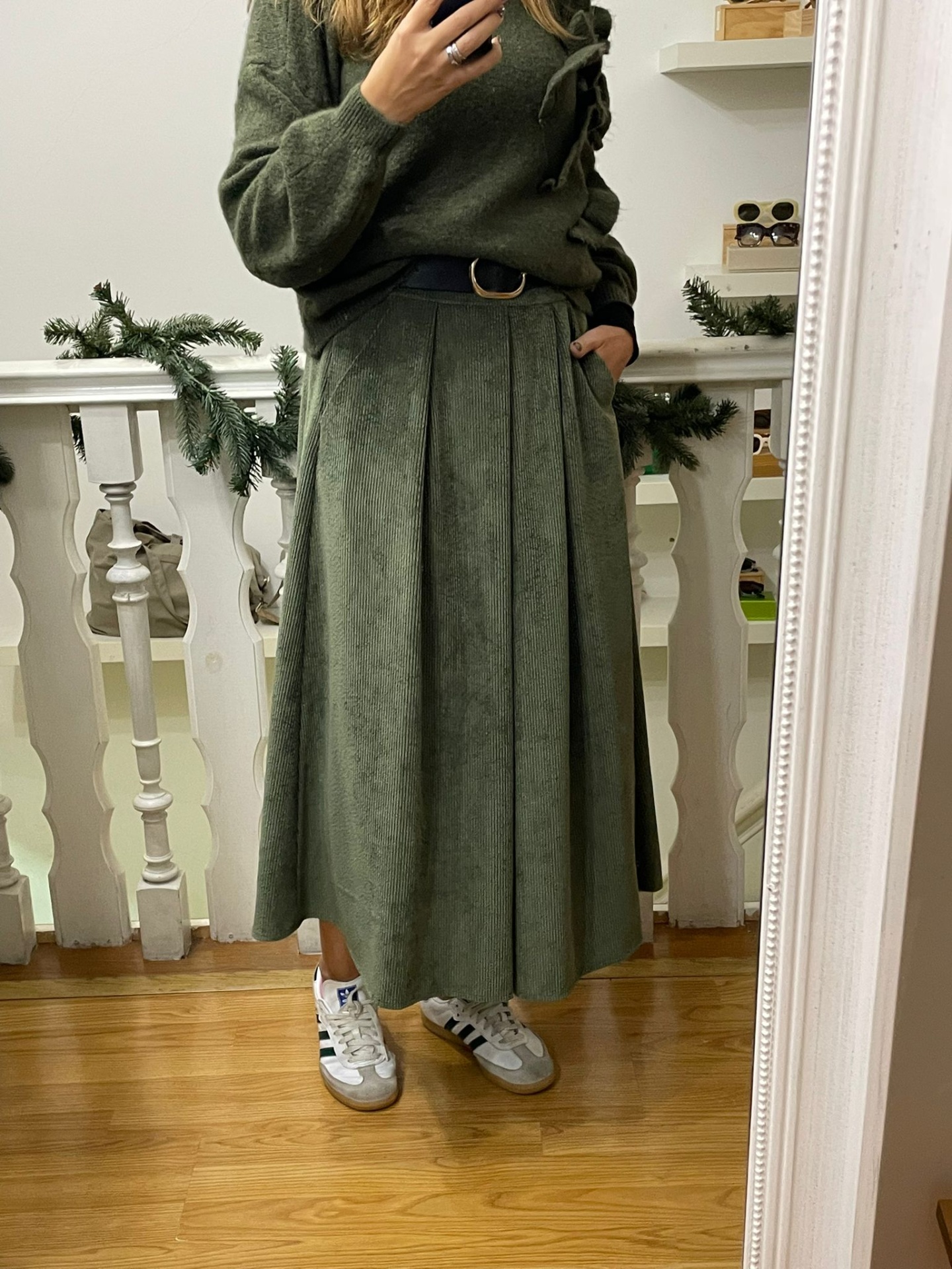 Mulher a usar saia midi verde, camisola verde de malha, cinto preto e sapatilhas brancas numa divisão com chão de madeira e decoração natalícia.