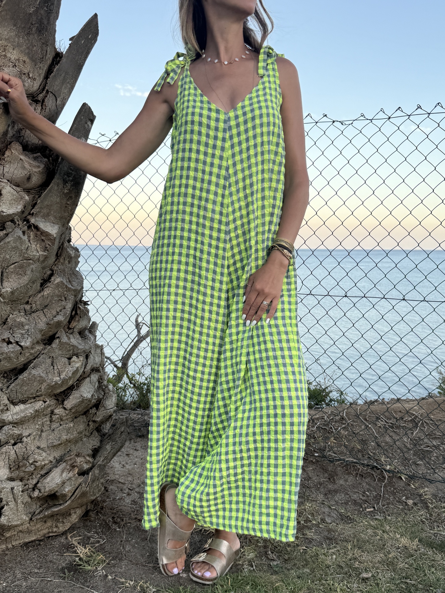 Vestido comprido xadrez verde lima e cinza com alças de laço junto a cerca e mar ao fundo