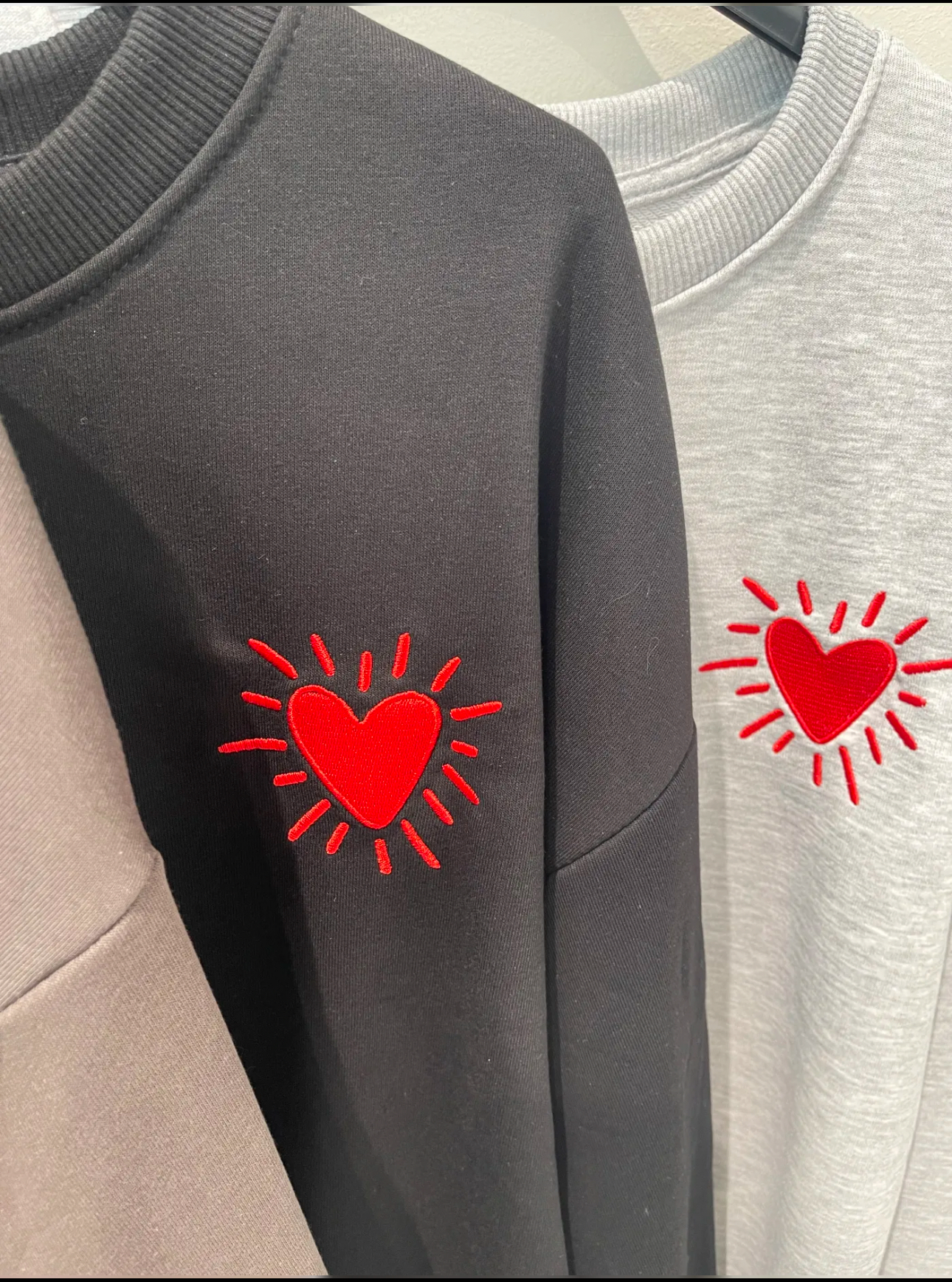 Três sweatshirts bege, preto e cinza claro com coração vermelho bordado no peito.