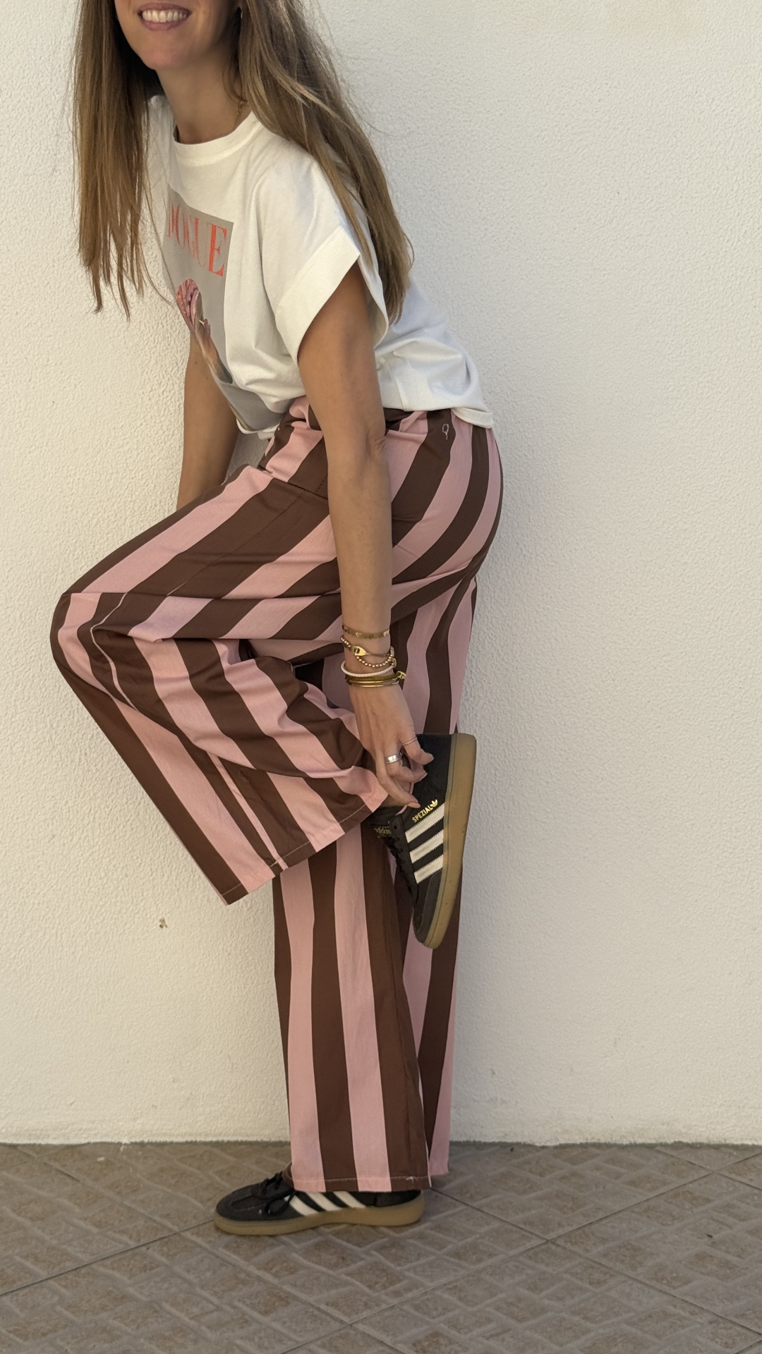 Calças largas riscas castanhas e rosa, t-shirt branca com impressão, sapatilhas desportivas pretas e brancas, parede branca, chão azulejo.