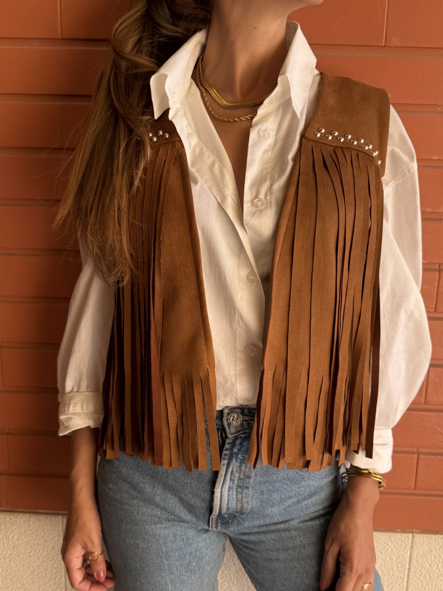 Colete castanho de camurça com franjas e tachas sobre camisa branca e calças de ganga