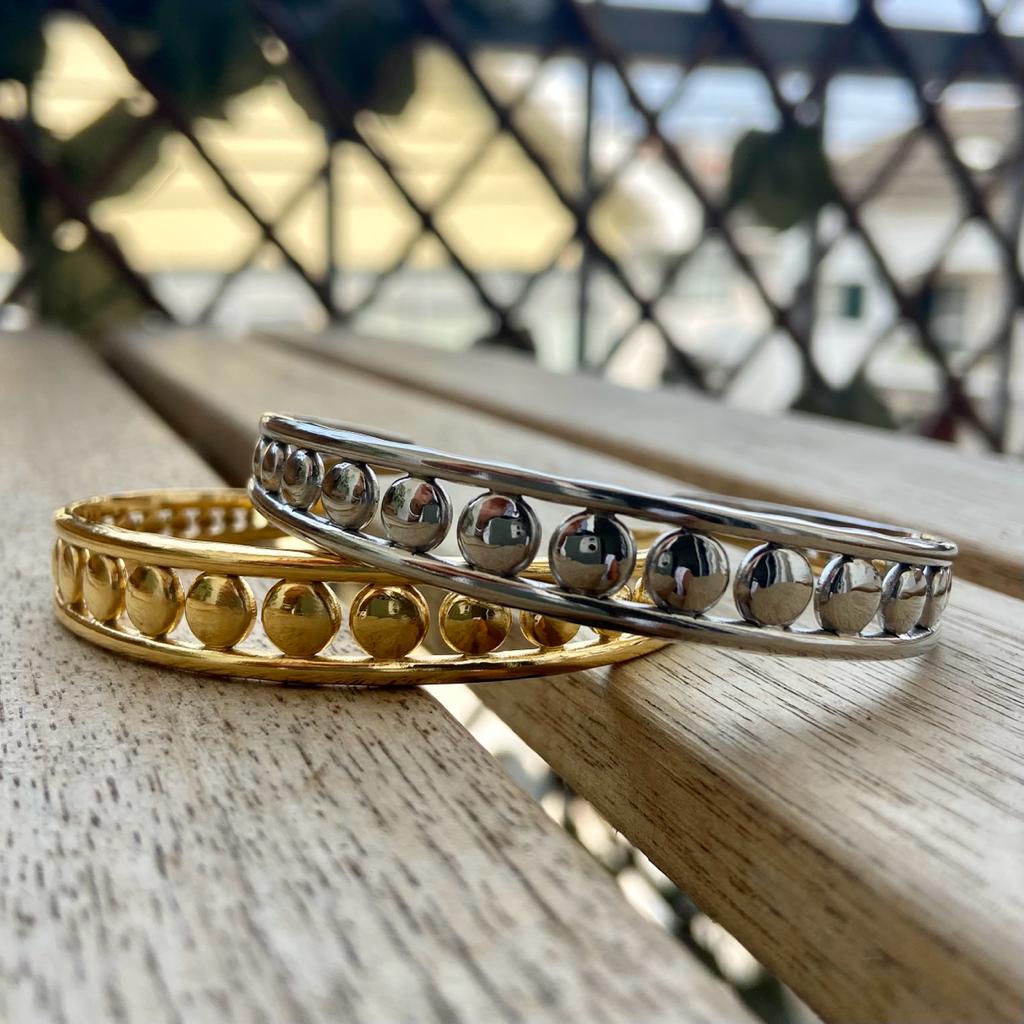 Dois braceletes metálicos dourado e prata com padrão de bolas colocados sobre mesa de madeira.