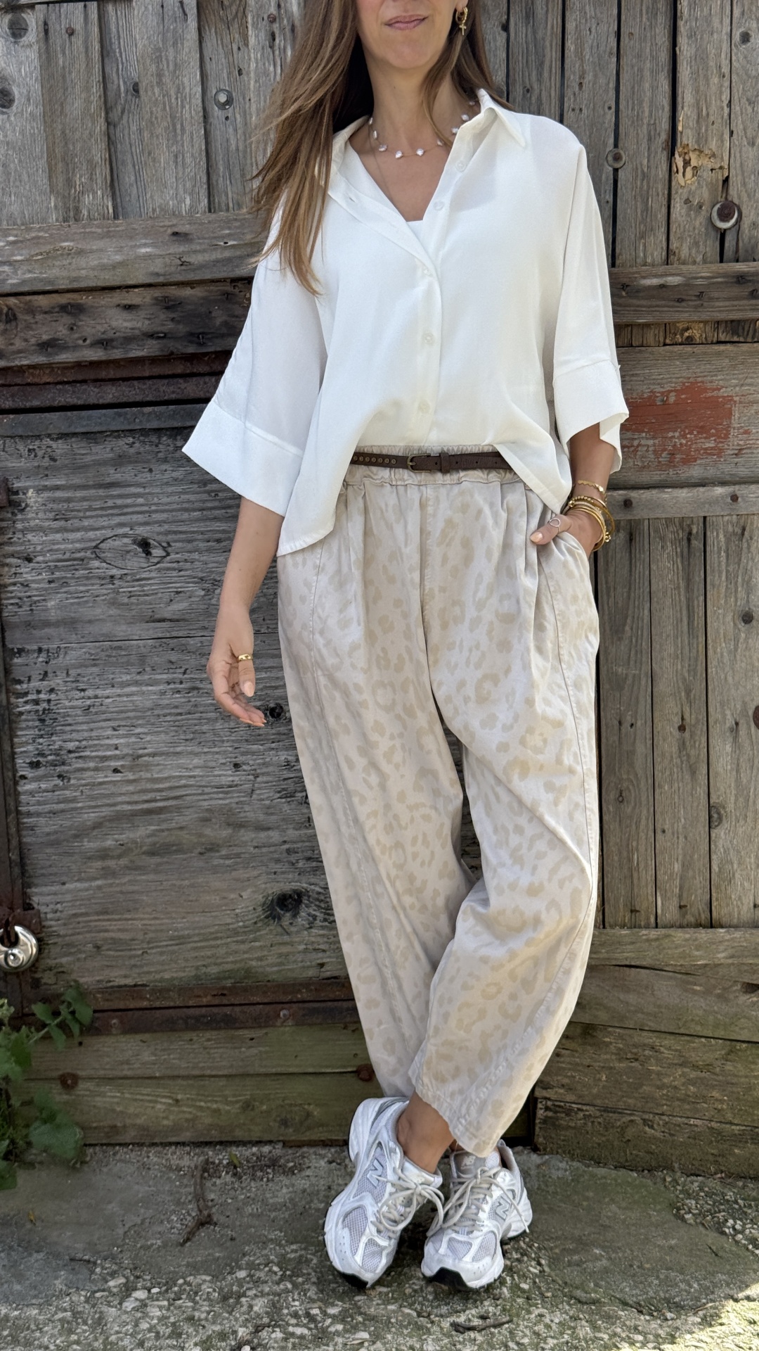 Mulher a usar camisa branca e calças largas bege com padrão animal print, ténis brancos, com madeira rústica ao fundo.