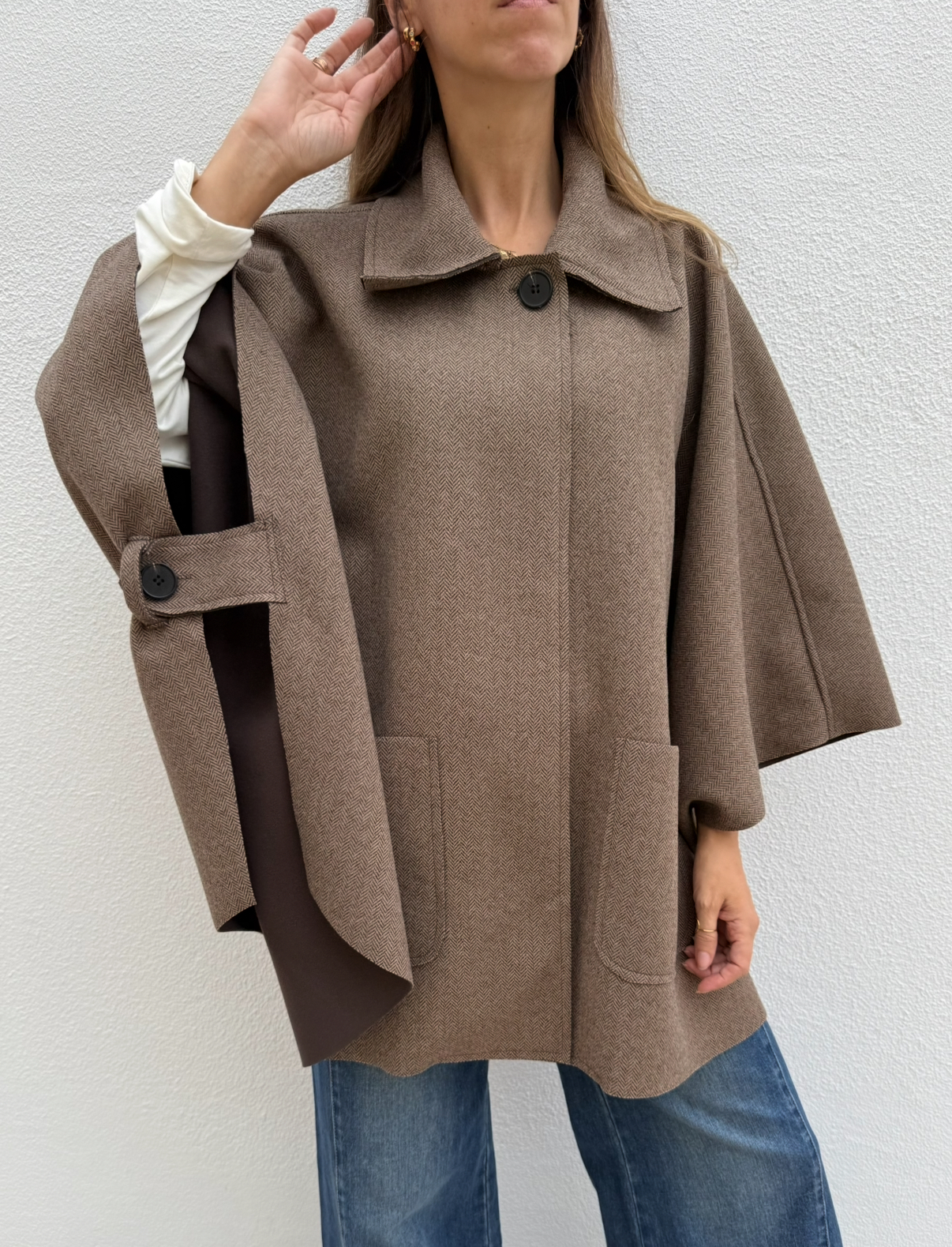 Capa castanha com padrão espinha de peixe e botões, vista parcial da parte superior de pessoa com camisa branca e jeans.