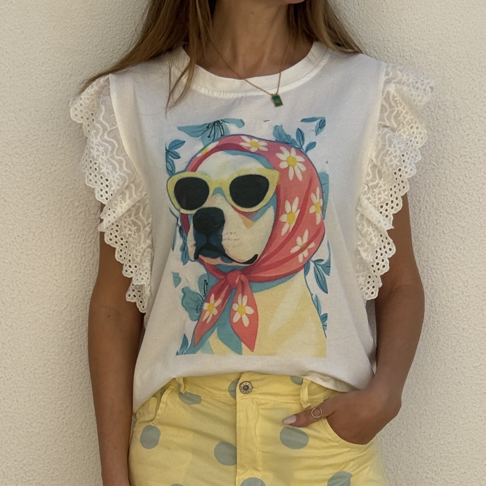 T-shirt branca com estampa de cão com óculos de sol e lenço, mangas rendadas e calças amarelas com pontos cinzentos.