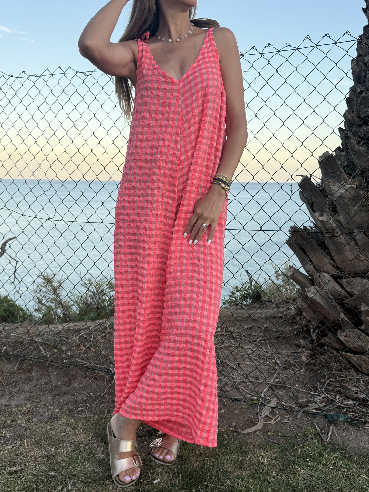 Vestido comprido xadrez vermelho e rosa com alças finas e sandálias metálicas douradas ao ar livre junto ao mar.