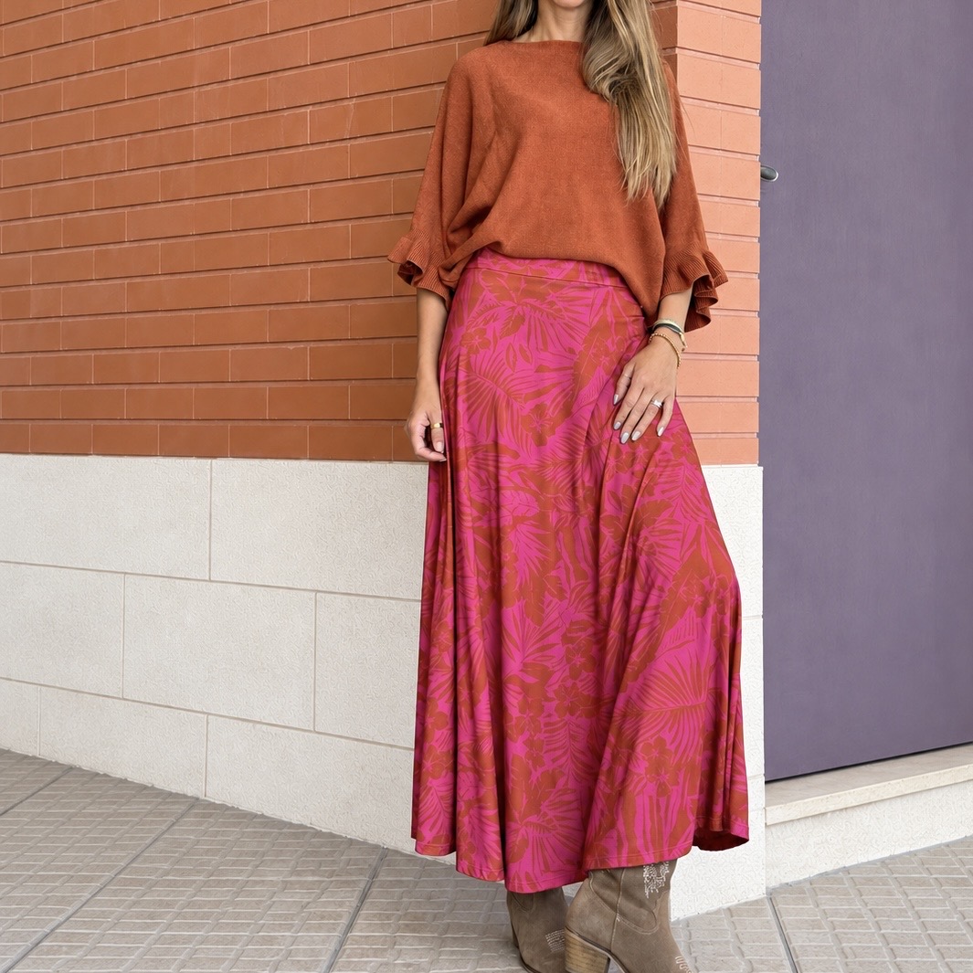 Mulher com saia longa rosa com padrão floral castanho e suéter castanho à frente de parede de tijolo e porta roxa