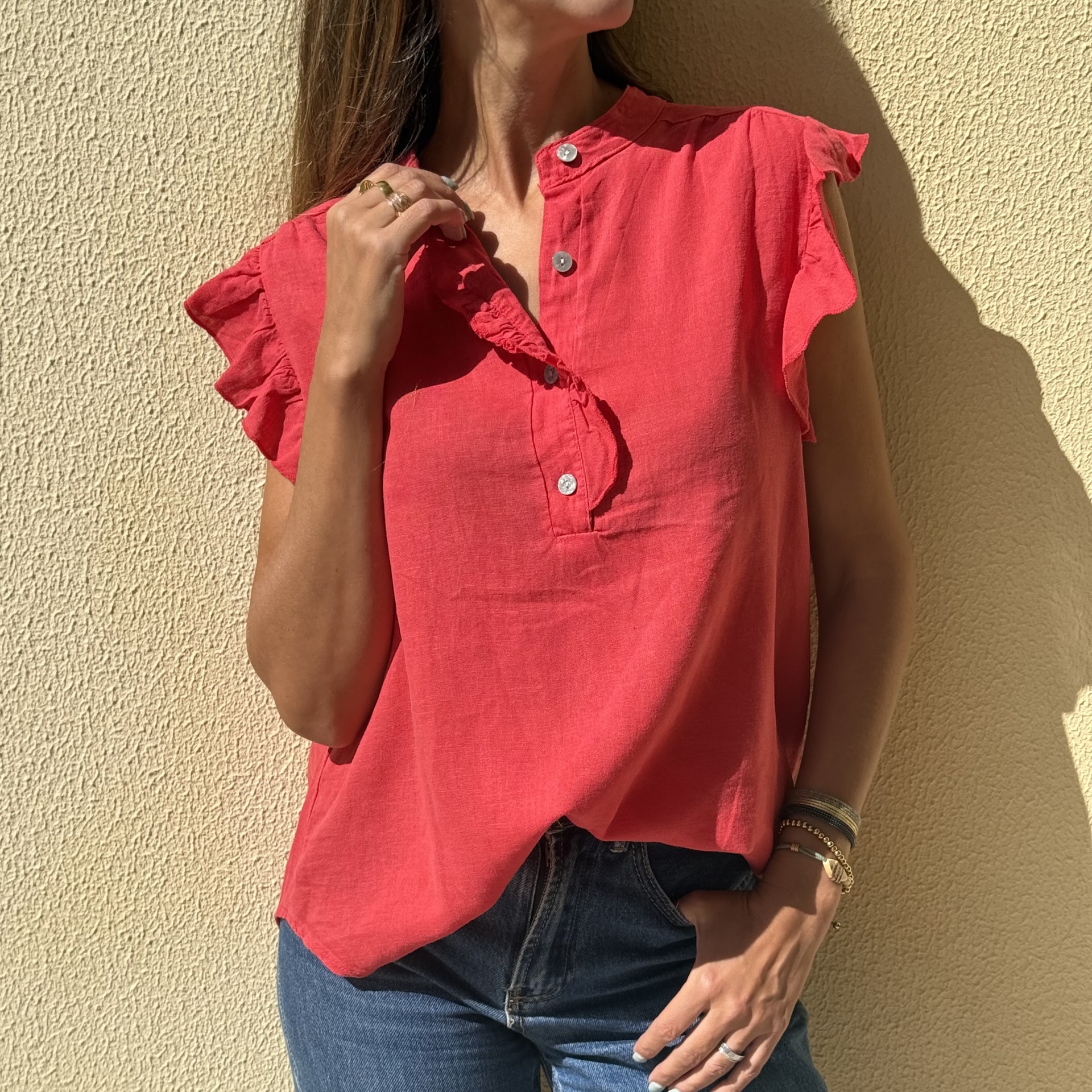 Blusa coral com botões e folhos nas mangas, calças de ganga, acessórios de joalharia e parede amarela texturada em fundo.