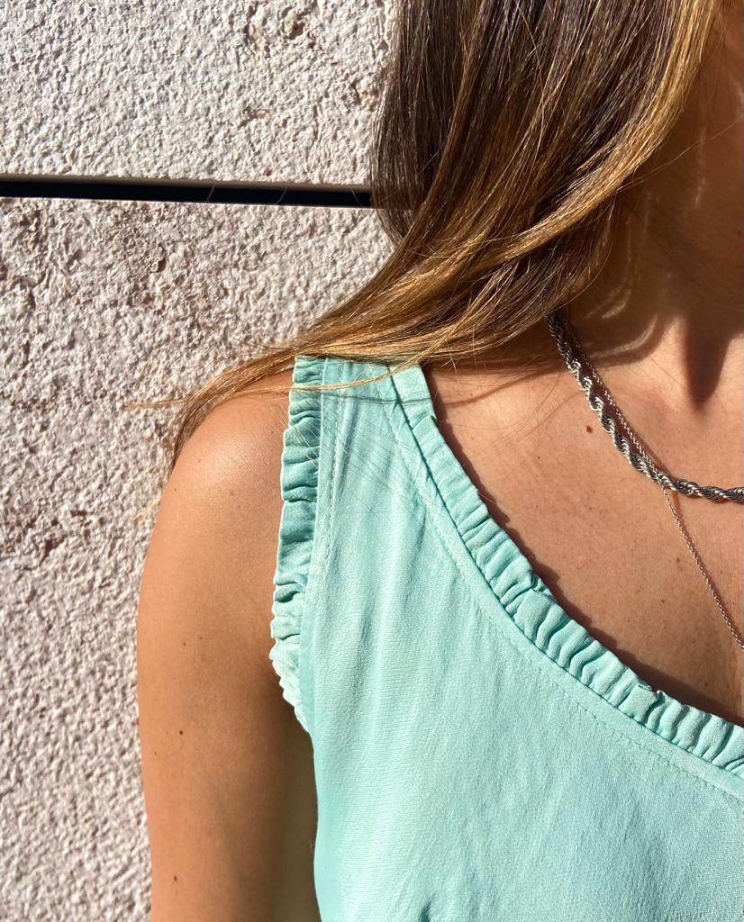 Blusa azul claro com folhos no decote e alças, usada perto de uma parede clara.