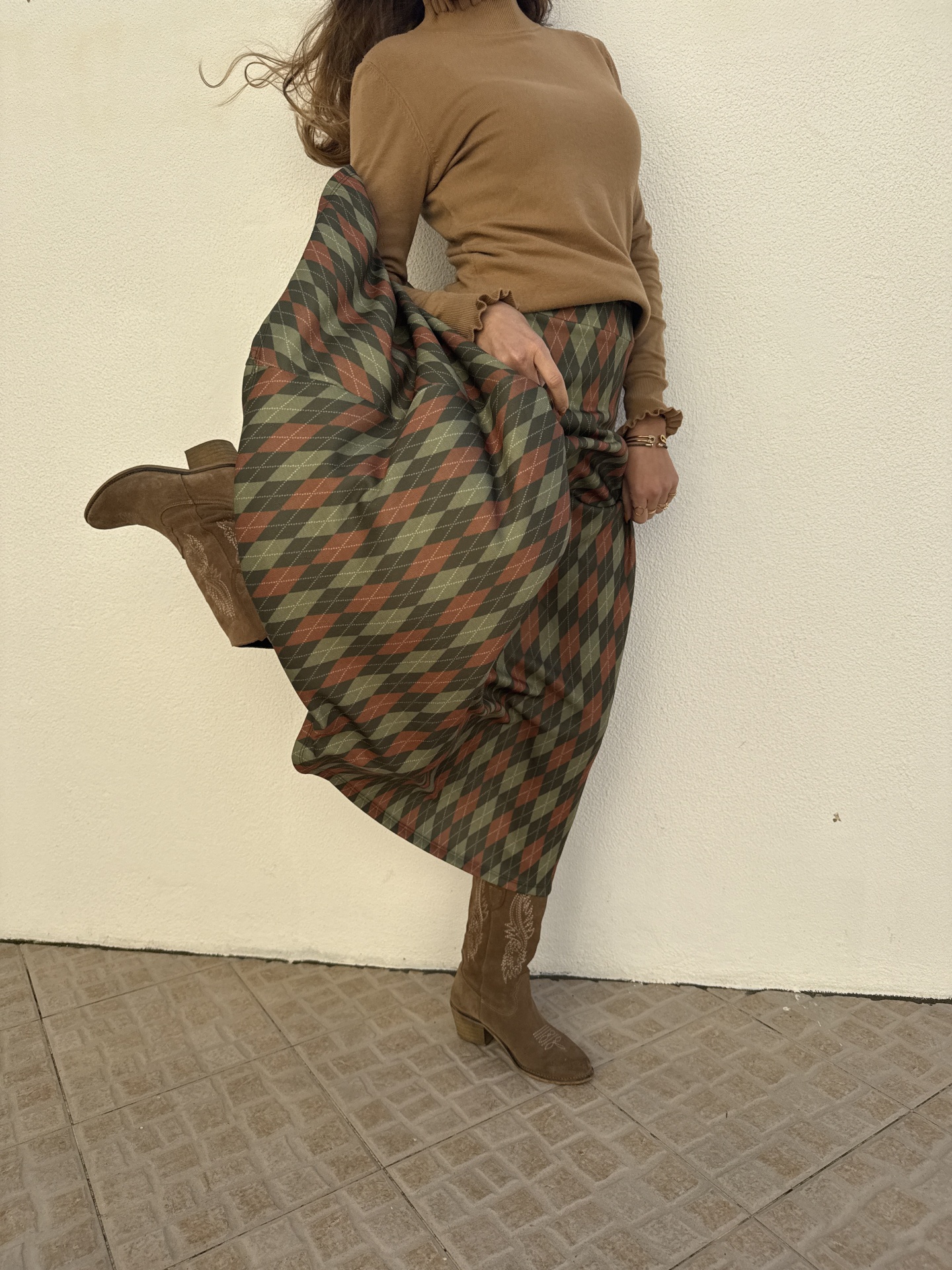 Saia midi com padrão argyle em tons de verde, castanho e bege, com camisola castanha e botas castanhas de cano alto contra parede branca