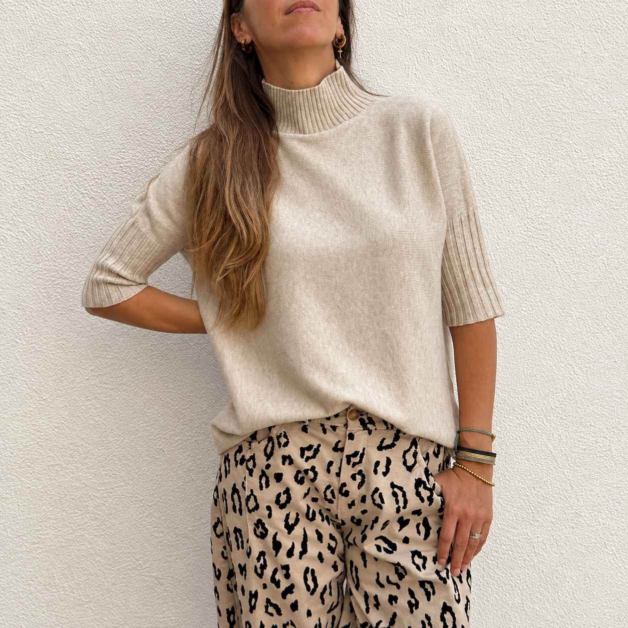 Mulher com camisola bege de malha e calças bege com padrão animal, pulseiras no pulso direito.