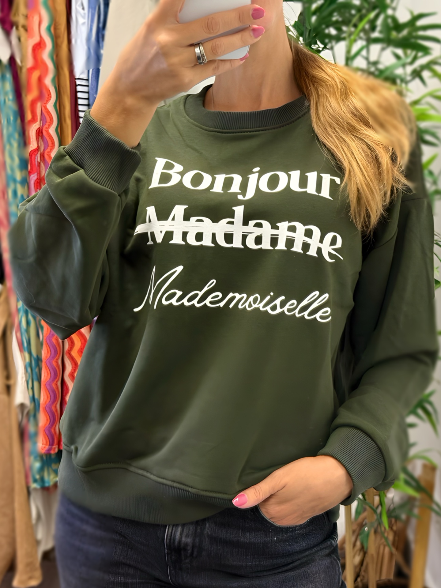 Pessoa com sweatshirt verde escuro com texto branco em ambiente interior com plantas e roupas ao fundo