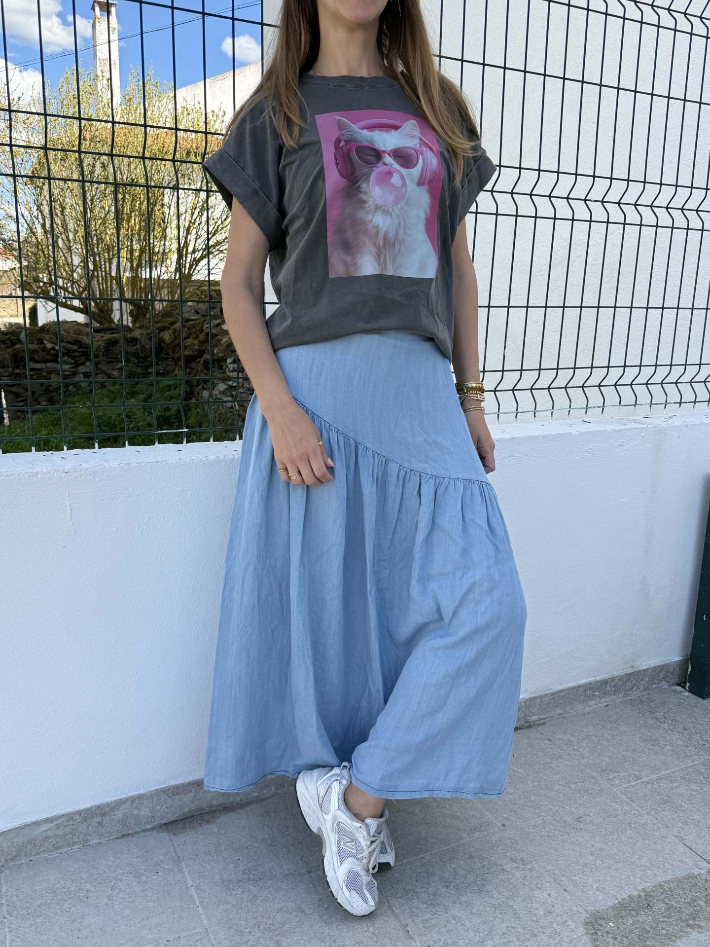 Mulher com camiseta cinza escuro com estampa de gato, saia azul comprida e sapatilhas brancas ao ar livre