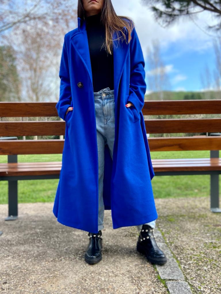 Mulher usando casaco azul royal comprido, jeans claros e botas pretas perto de banco de parque