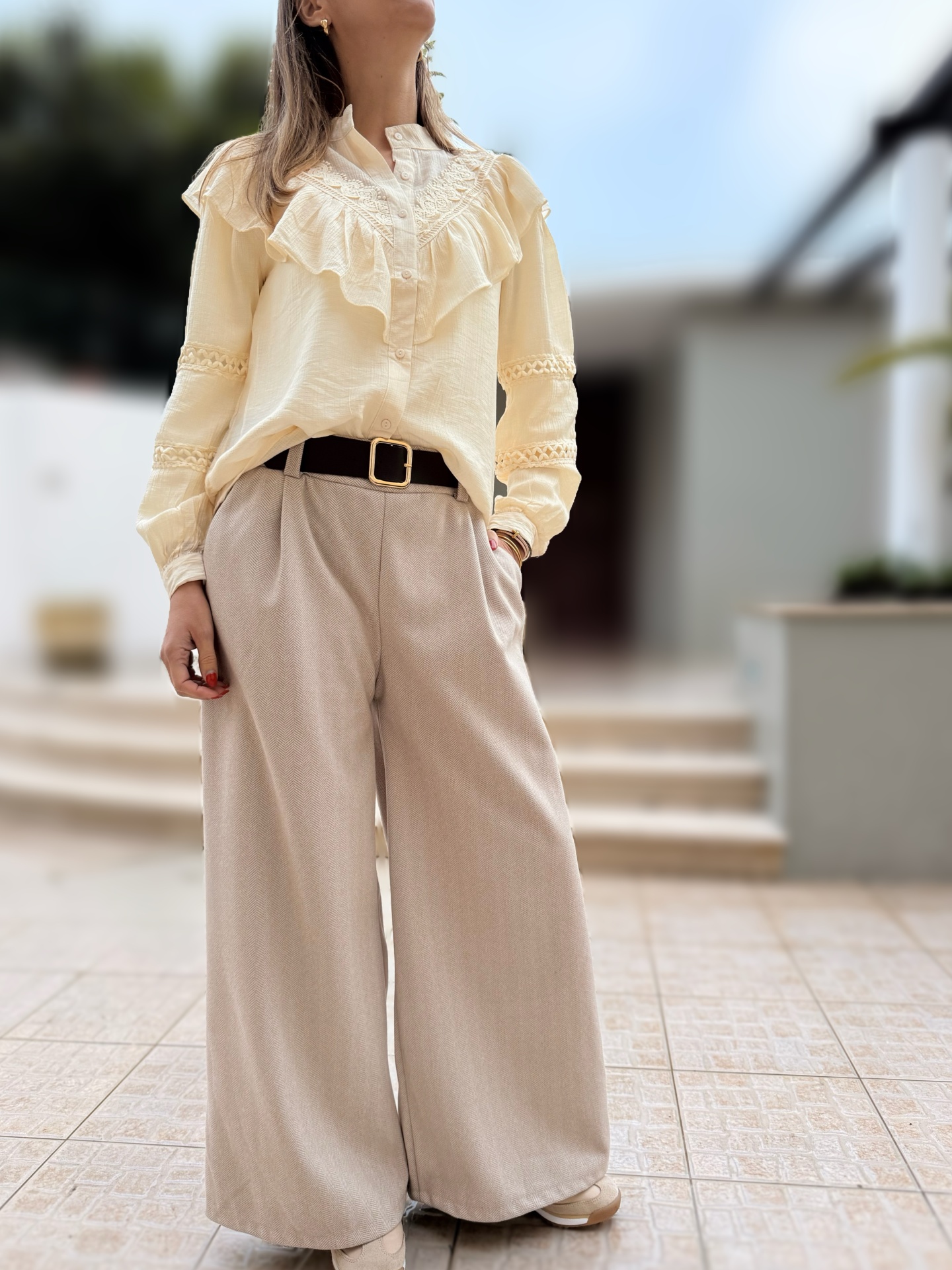 Pessoa de pé vestindo camisa bege com folhos e calças largas cinza claro com cinto preto.