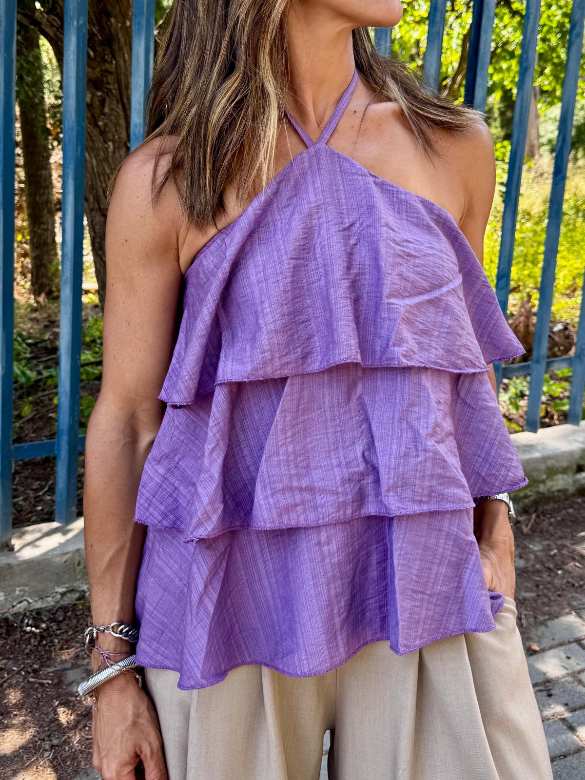 Top lilás com camadas e calças bege em ambiente exterior com cerca azul ao fundo.
