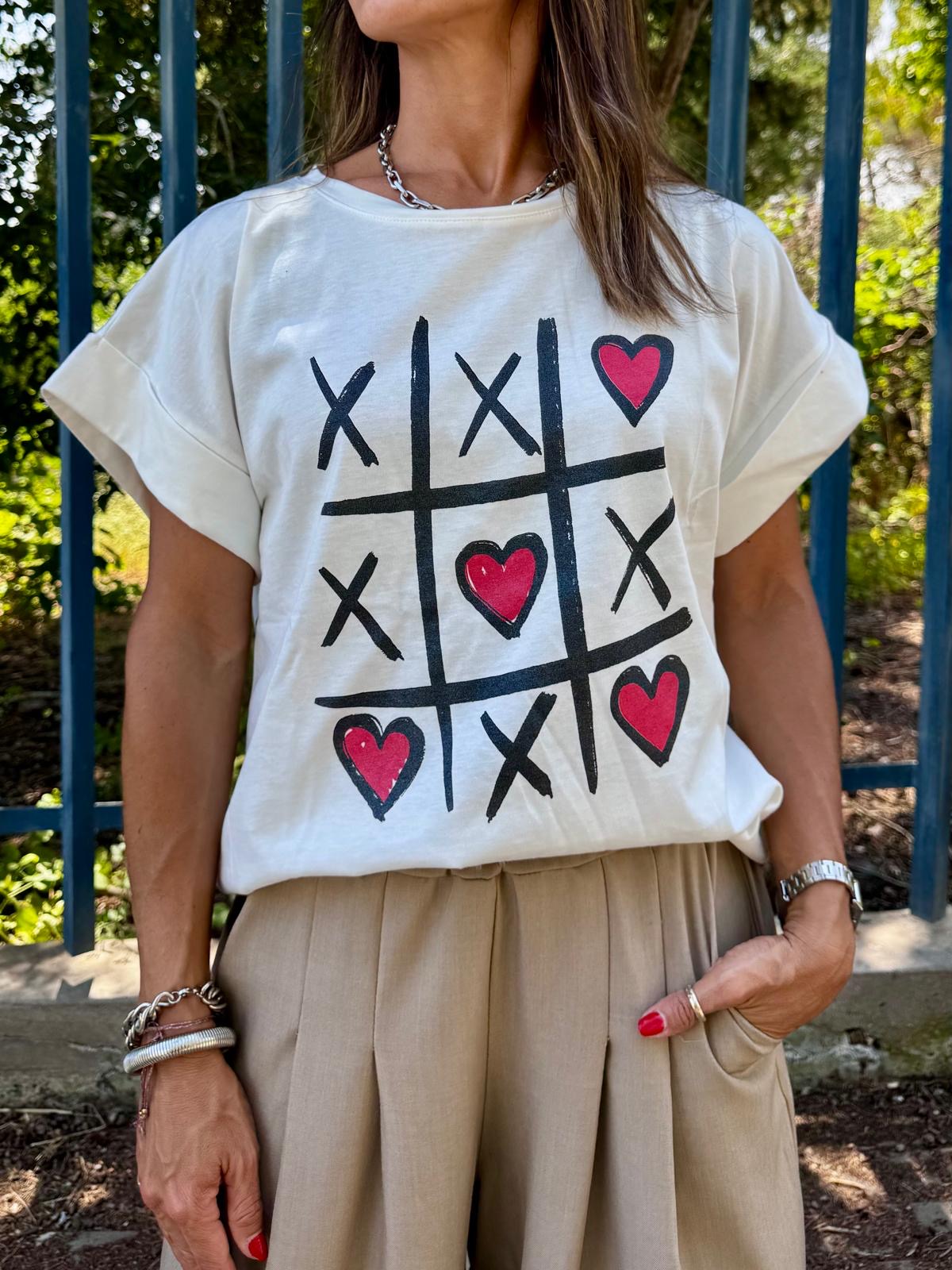 T-shirt branca com desenho de jogo-da-velha com corações vermelhos e calças beje ao ar livre