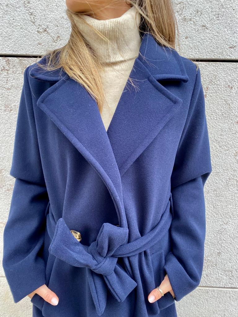 Casaco azul escuro com cinto e gola larga sobre camisola creme junto a parede clara