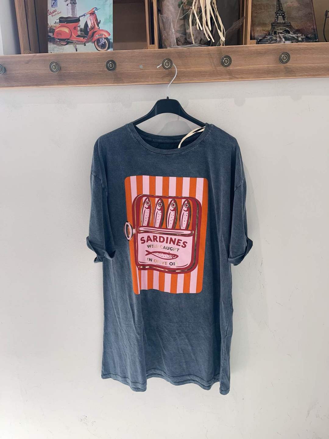 T-shirt cinza escuro com estampa de lata de sardinhas e texto em inglês pendurada na parede