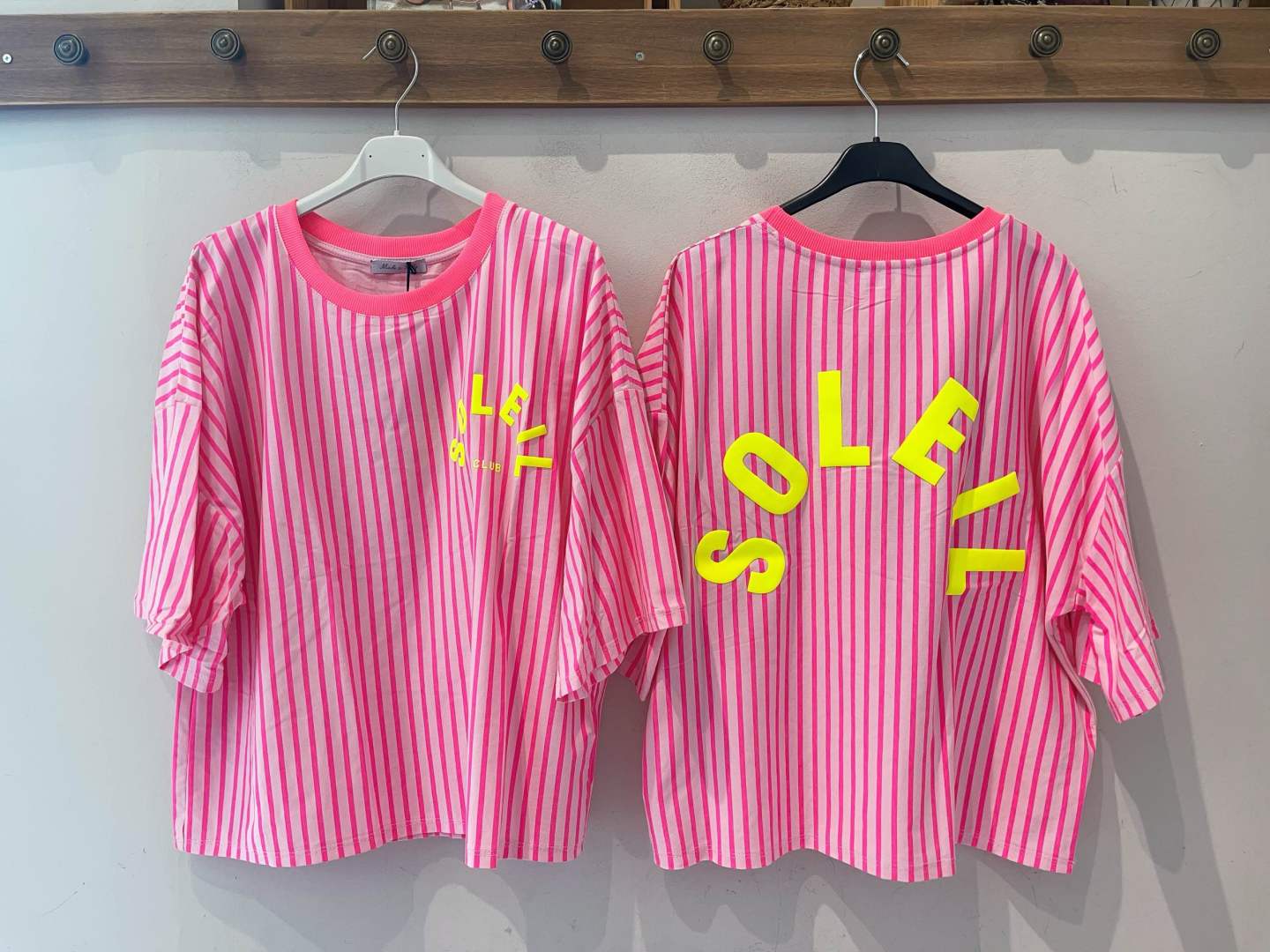 Duas t-shirts rosa às riscas vermelhas com texto amarelo penduradas num cabide