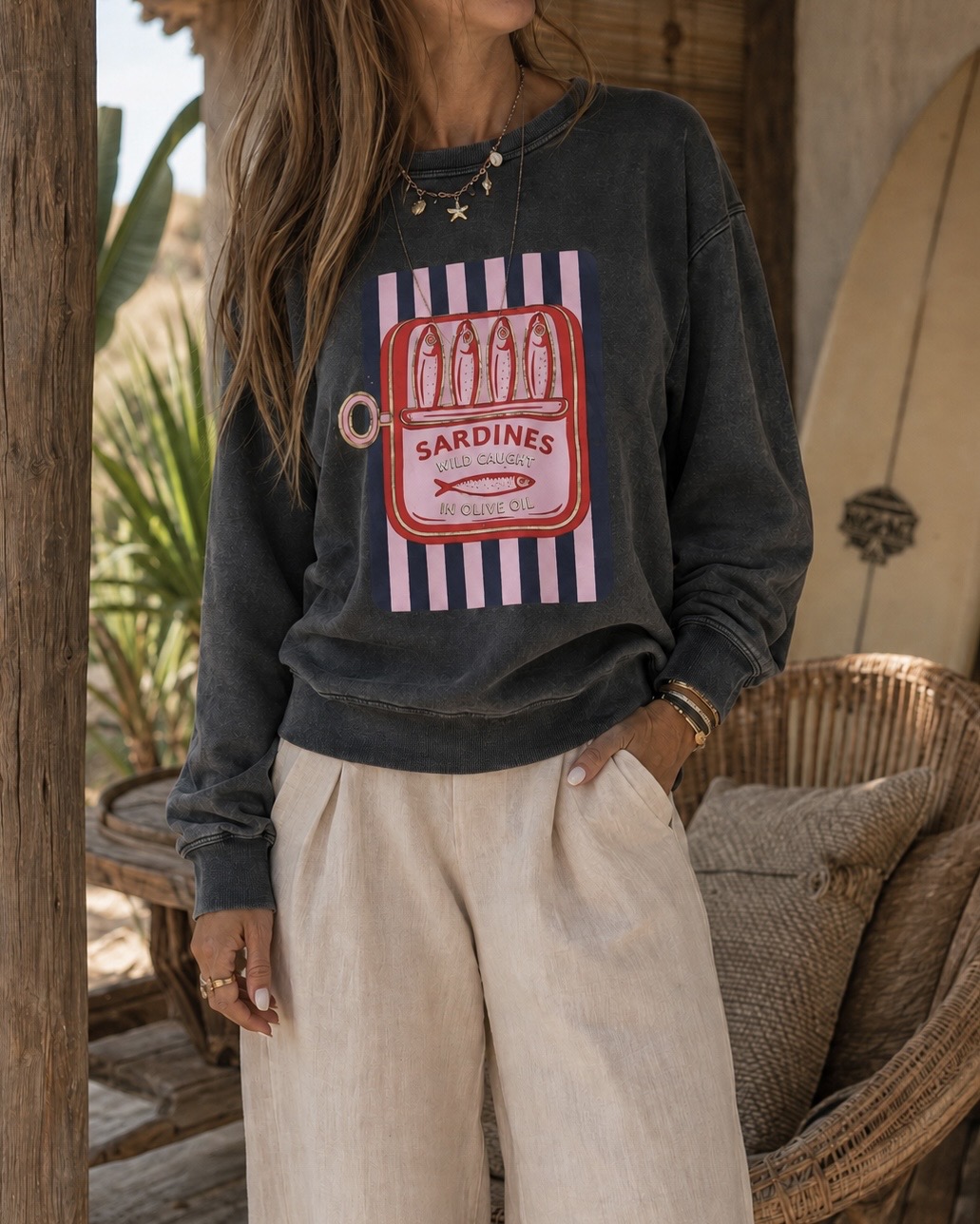 Camisola cinza escura com estampa colorida de lata de sardinhas e calças largas bege, em ambiente rústico com cadeira de vime e planta.