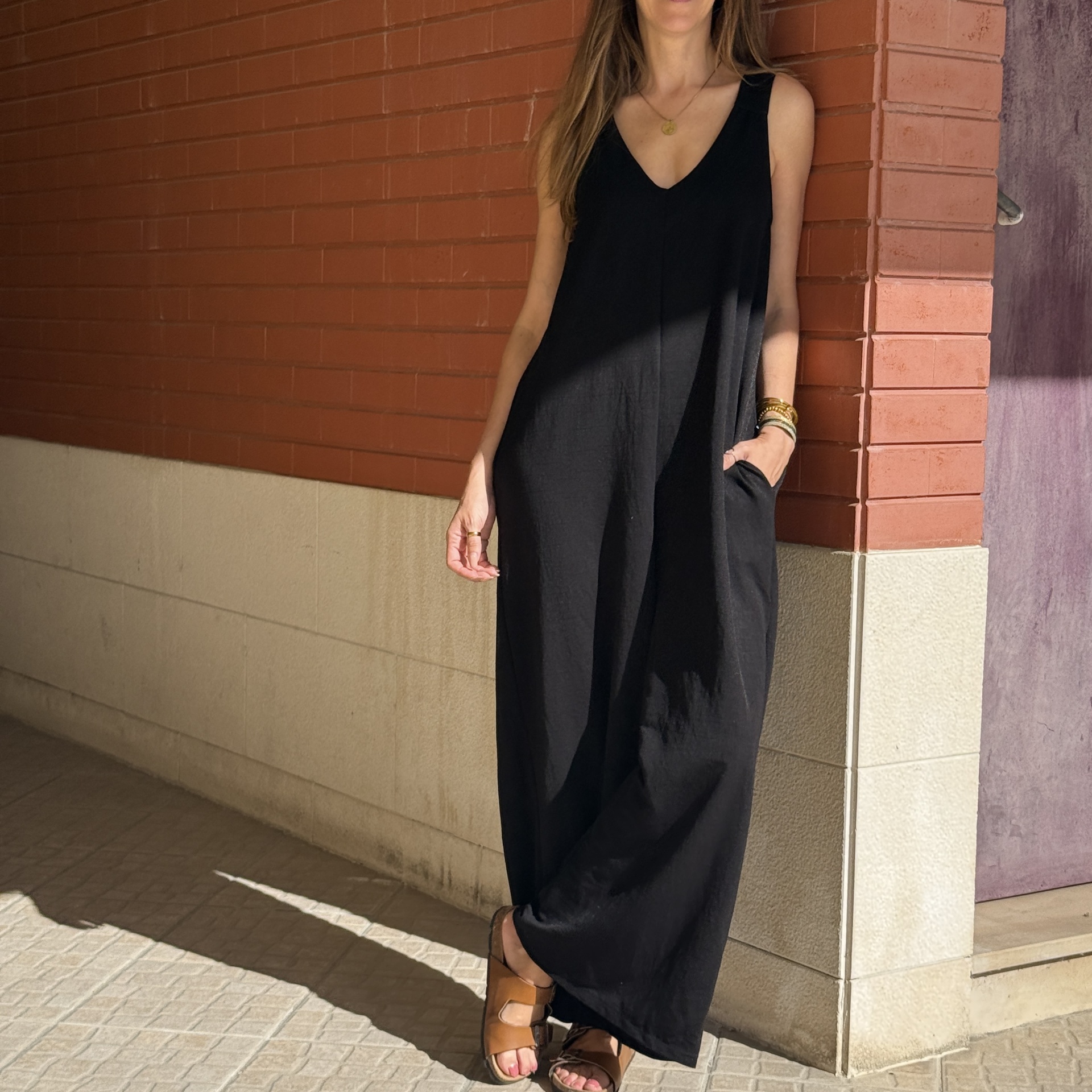 Vestido longo preto com sandálias marrons e parede de tijolos vermelhos ao fundo
