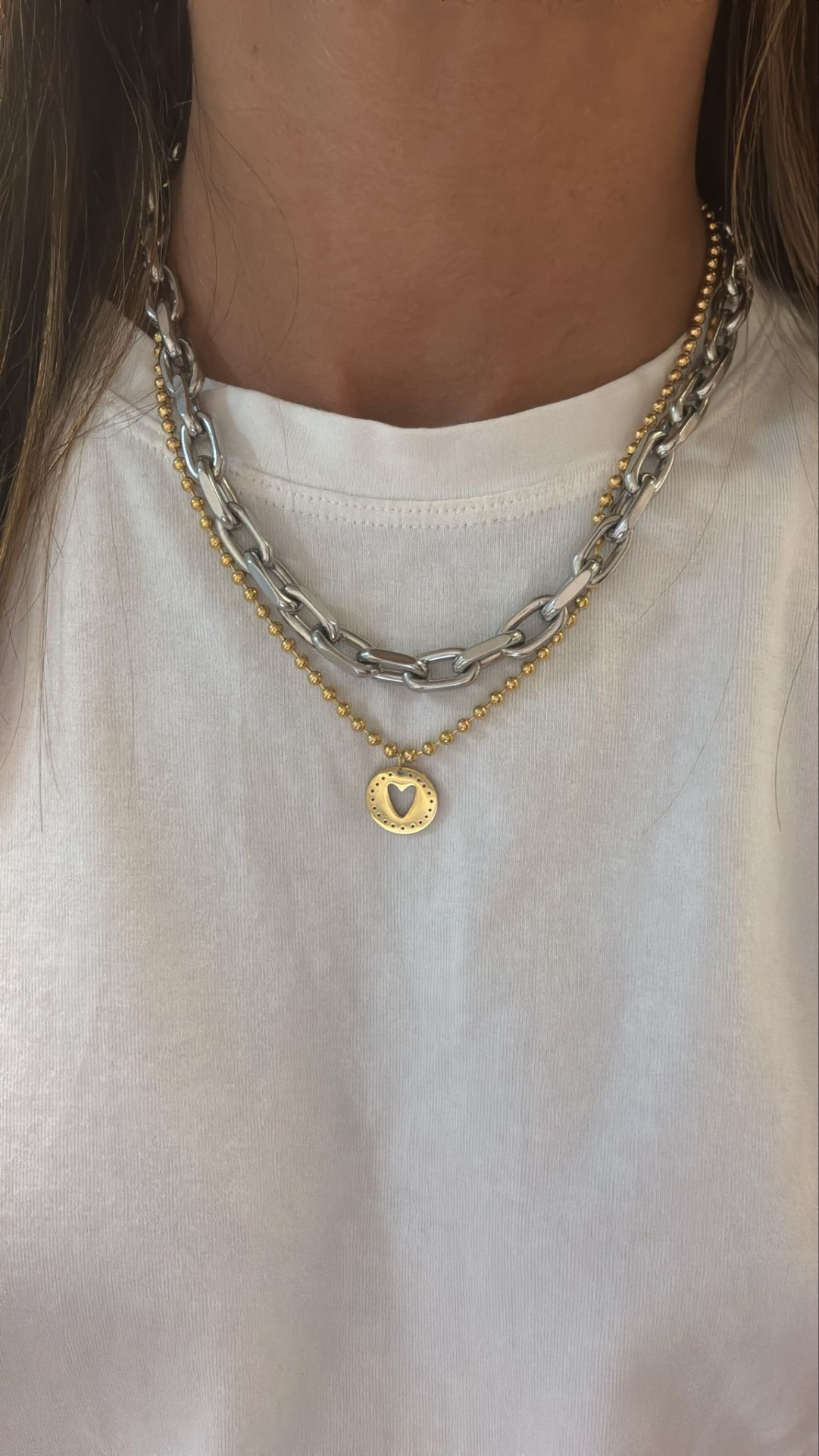 Colares prateado de elos grandes e colar dourado com bolinhas e pendente em forma de coração sobre t-shirt branca