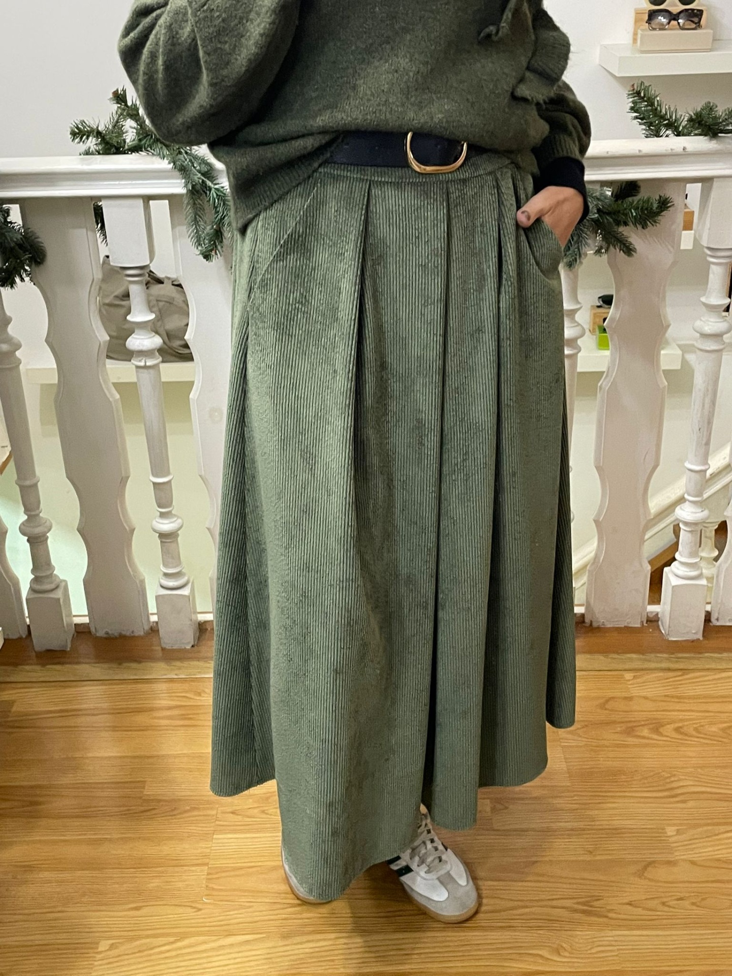 Saia longa verde com pregas e cinto preto, usada com camisola verde e sapatilhas brancas, junto a grade branca decorada para Natal