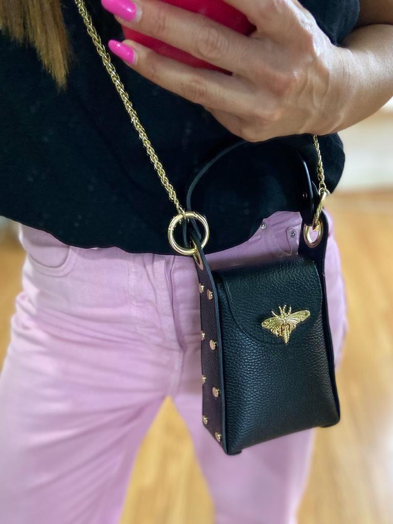 Bolsa preta pequena com abelha dourada e corrente dourada, vestida por pessoa com unhas rosa e calças rosas