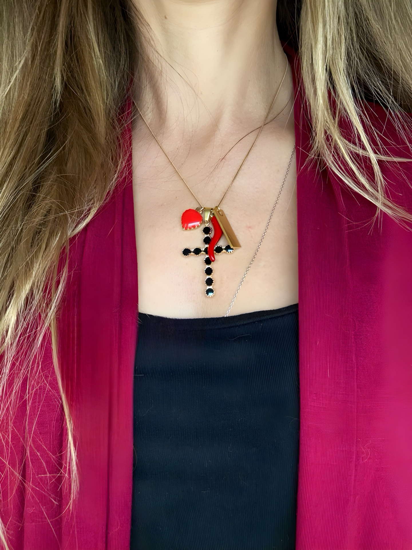 Colar dourado com cruz preta, corno vermelho, coração vermelho e pendente retangular, em mulher com casaco vermelho e top preto.